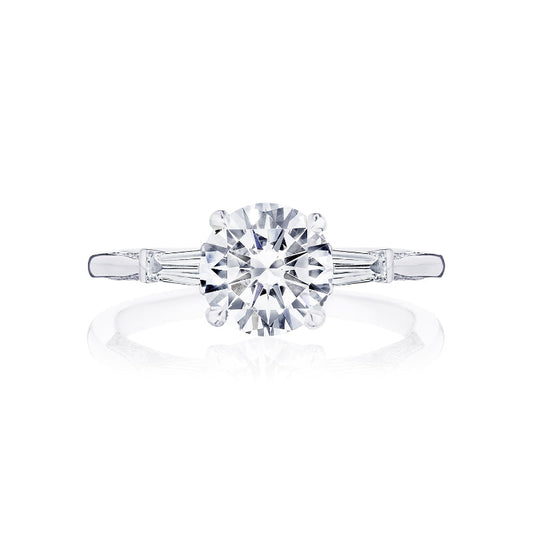An 18K white gold engagement ring by Tacori, featuring a round brilliant center diamond accented with side stones and a diamond accent band. The elegant design echoes the classic Tacori crescent style, set against a plain white background.