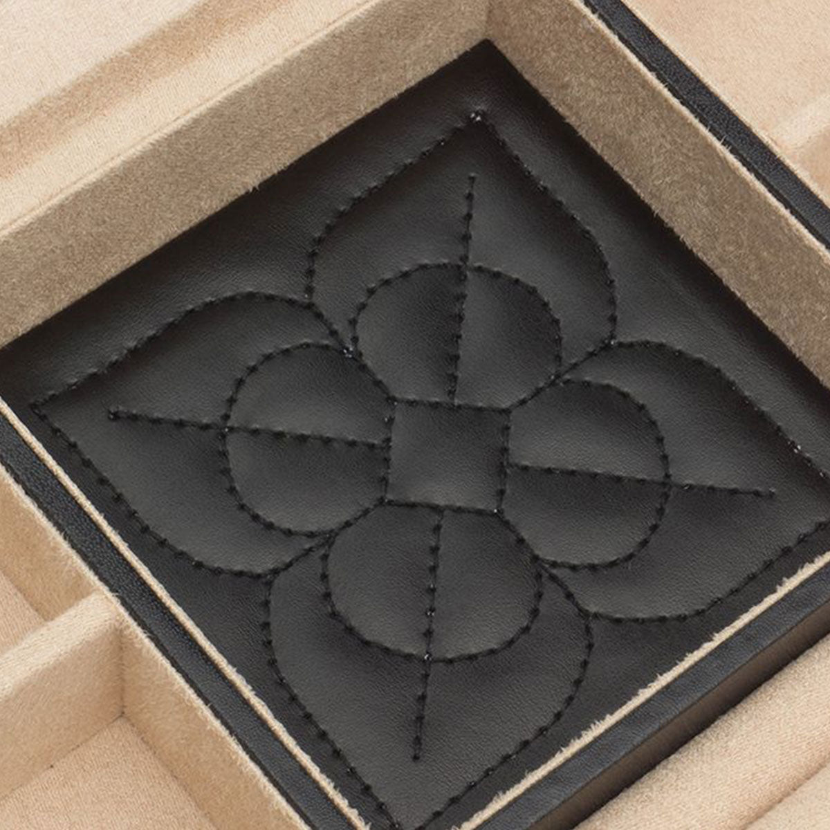 Close-up of a decorative black leather patch inside the WOLF 1834 Marrakesh Black Jewellery Box. The patch, featuring an embossed geometric flower design with stitched details, sits neatly in a square slot. The box's beige compartments are enhanced by the protective LusterLoc™ lining from WOLF.