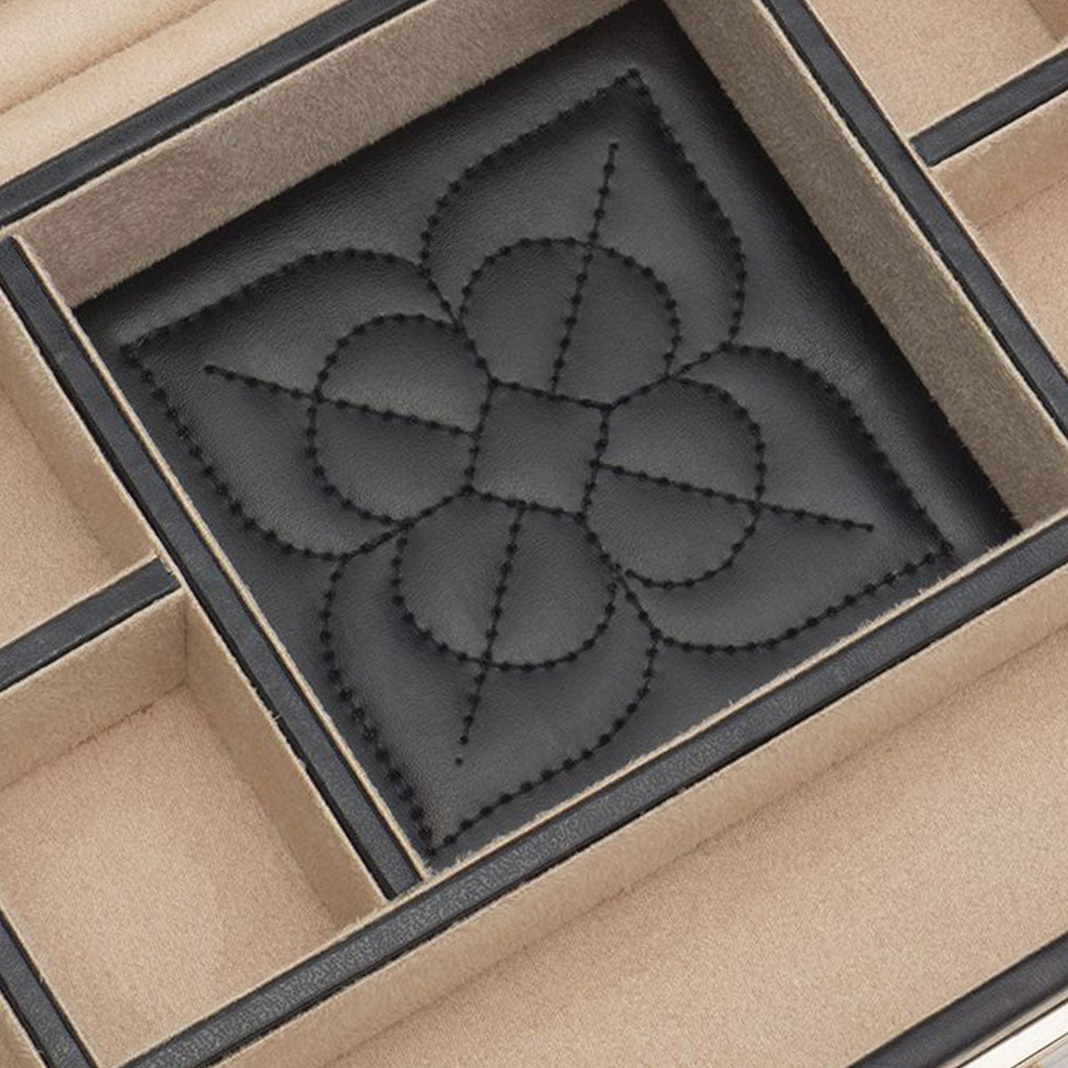 A close-up of the WOLF 1834 Marrakesh Flat Jewellery Box reveals a black embossed design reminiscent of a stylized flower or emblem, nestled inside its beige velvet-lined compartment. The leather jewelry box exudes elegance with divider sections and subtle gold studding.