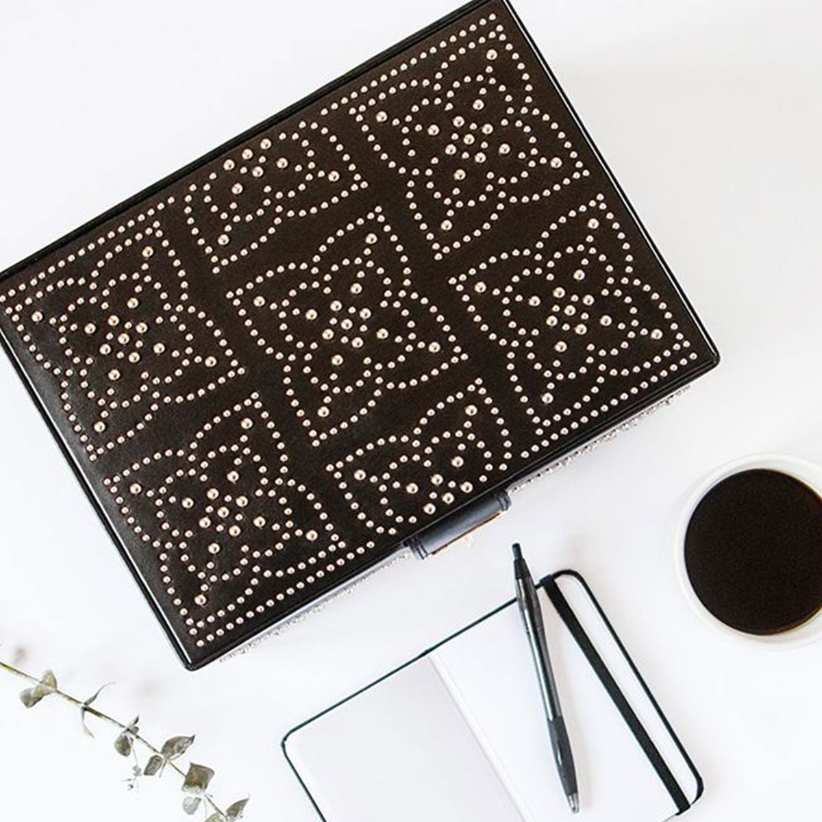 A WOLF 1834 Marrakesh Flat Jewellery Box with gold detailing and an intricate geometric design sits on a white surface next to an open notebook with a pen, a cup of black coffee, and a small green branch.