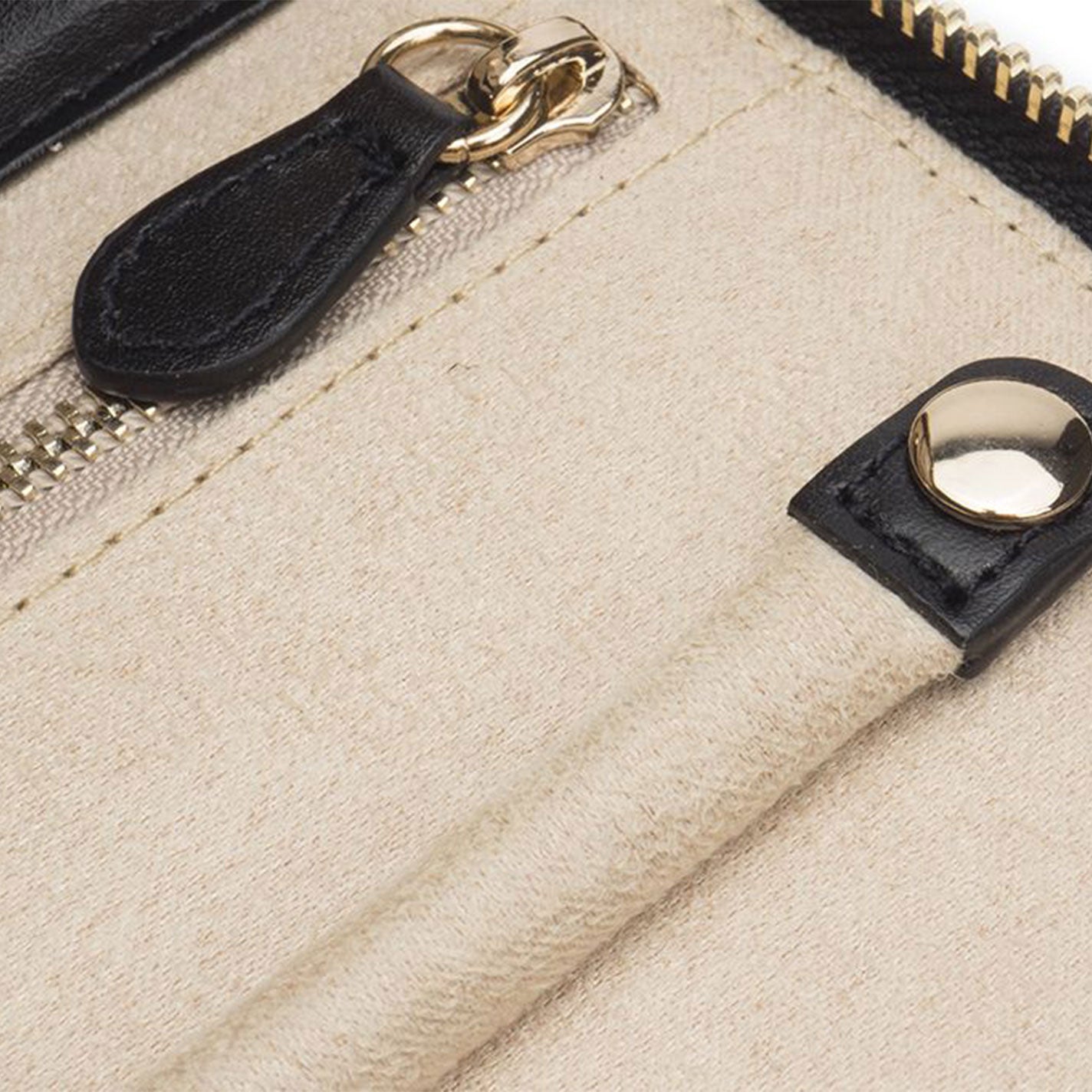 Close-up of a beige fabric WOLF 1834 Marrakesh Jewellery Box, featuring a black leather zipper pull and gold-tone zipper. A black leather strap is attached with a shiny gold rivet, providing contrast to the textured fabric. The image emphasizes the box's craftsmanship and details, enhanced with LusterLoc™ technology.