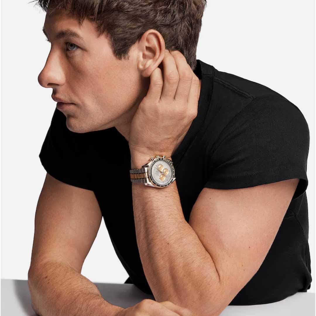 A person with short brown hair wearing a black t-shirt is sitting and resting his chin on his hand. He sports a stylish OMEGA Speedmaster Moonwatch Professional Co-Axial Master Chronometer Chronograph 42mm Watch, its metallic strap and round face with multiple chronograph dials standing out against the plain white background.