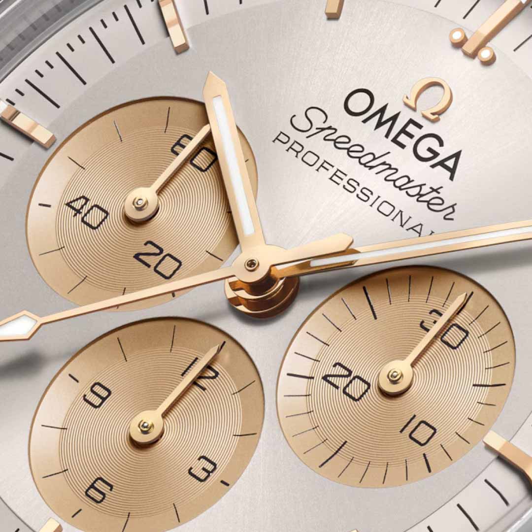 Close-up of a silver OMEGA Speedmaster Moonwatch Professional Co-Axial Master Chronometer Chronograph 42mm watch face with gold detailing. The hands are set to 10:10, showcasing the subdials and chronograph functionality, further accentuated by the textured design and OMEGA logo.