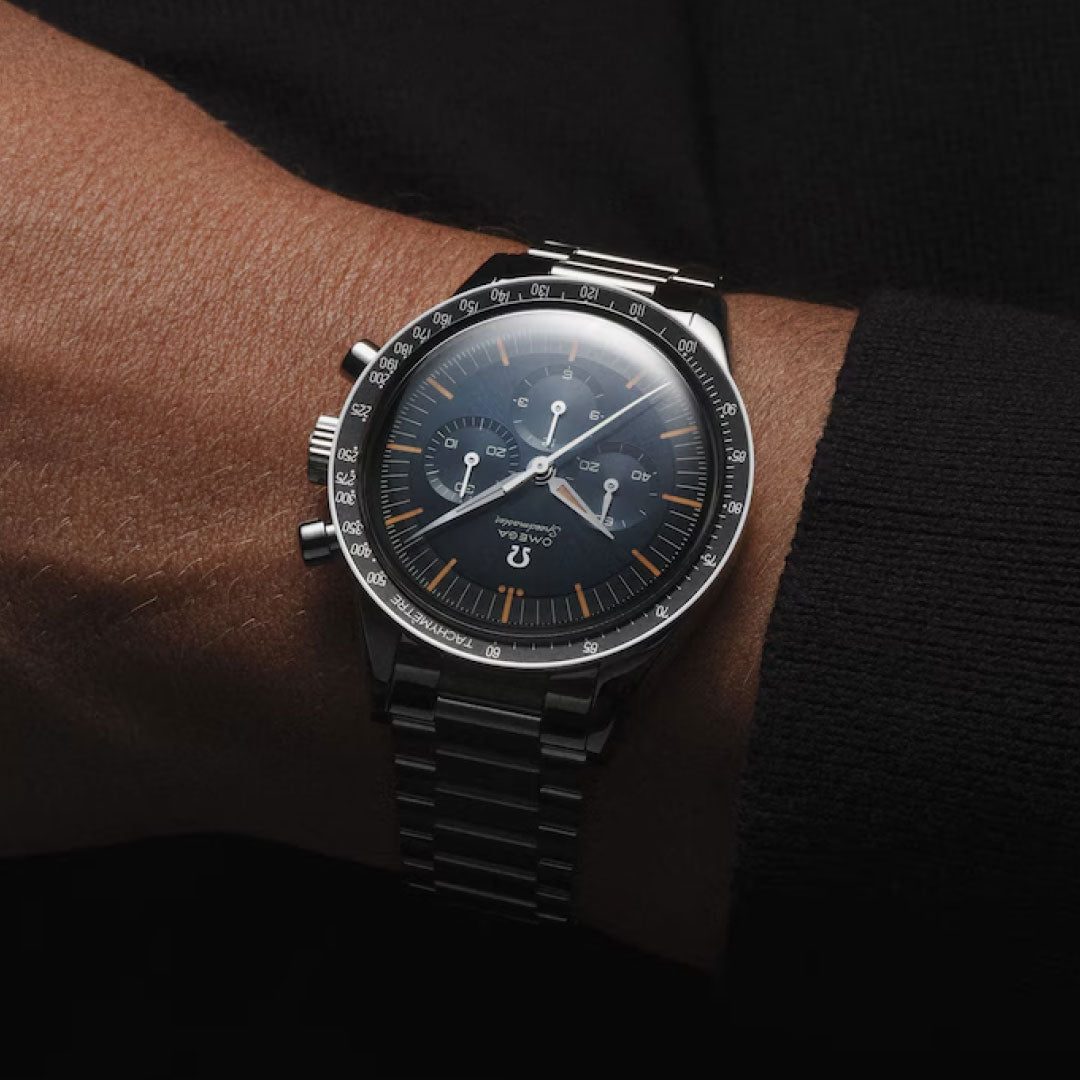 A close-up of a wrist wearing the OMEGA Speedmaster First OMEGA in Space Co-Axial Master Chronometer 39.7mm, featuring a black dial, white markings, tachymeter scale, and stainless steel case against a dark long sleeve.