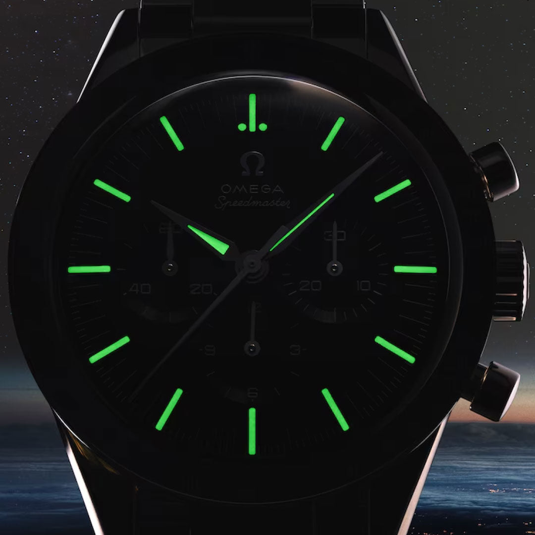Close-up of the OMEGA Speedmaster First OMEGA in Space Co-Axial Master Chronometer 39.7mm watch in the dark, green luminescent hour markers and hands glowing on its black dial, stainless steel case shining with a subtle space-themed backdrop.