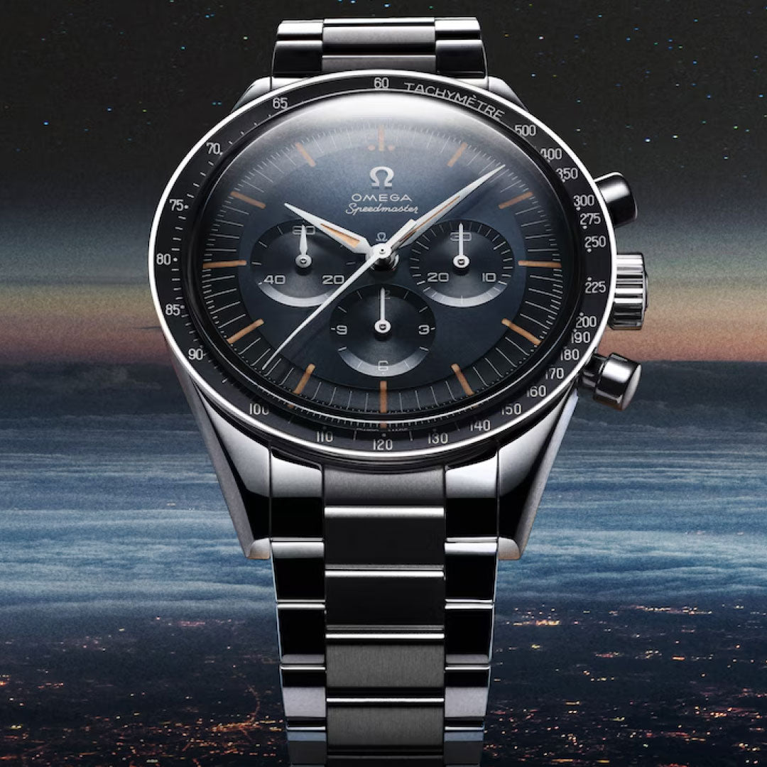 The OMEGA Speedmaster First OMEGA in Space Co-Axial Master Chronometer 39.7mm watch, featuring a black dial with three subdials and a tachymeter, is displayed above a city at night under a sky fading from blue to black.
