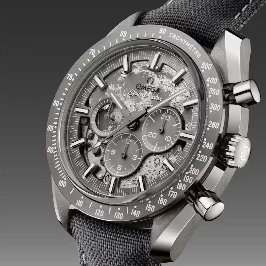 A close-up of the OMEGA Speedmaster Dark Side of the Moon Co-Axial Master Chronometer 44.25mm watch featuring chronograph subdials, a tachymeter bezel, and a fabric strap, set against a gradient gray background.