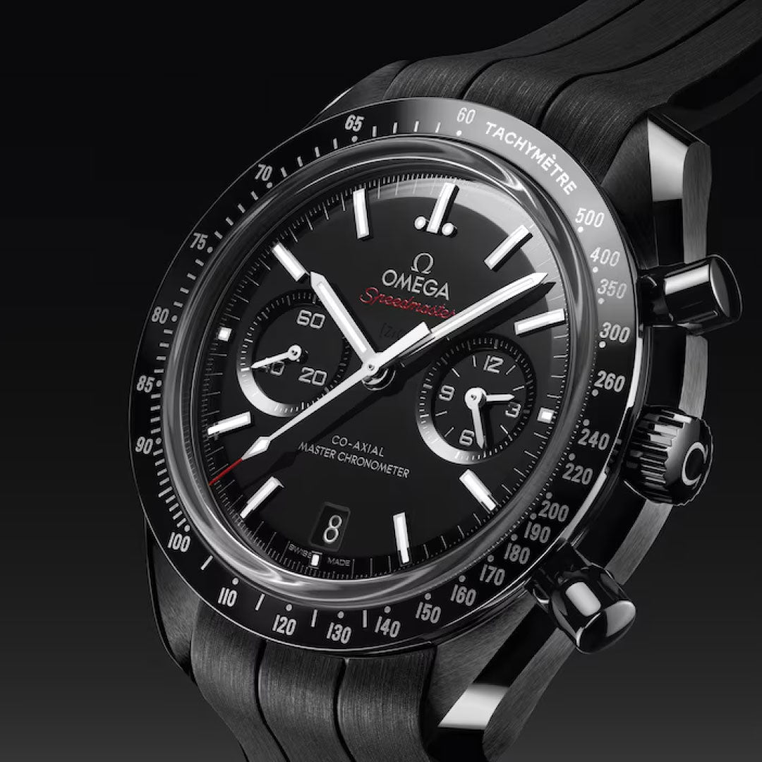 A close-up of the OMEGA Speedmaster Dark Side of the Moon Co-Axial Master Chronometer 44.25mm watch shows its black ceramic case, three subdials, tachymeter bezel, white hands and markers, red details, and a black strap on a dark gradient backdrop.