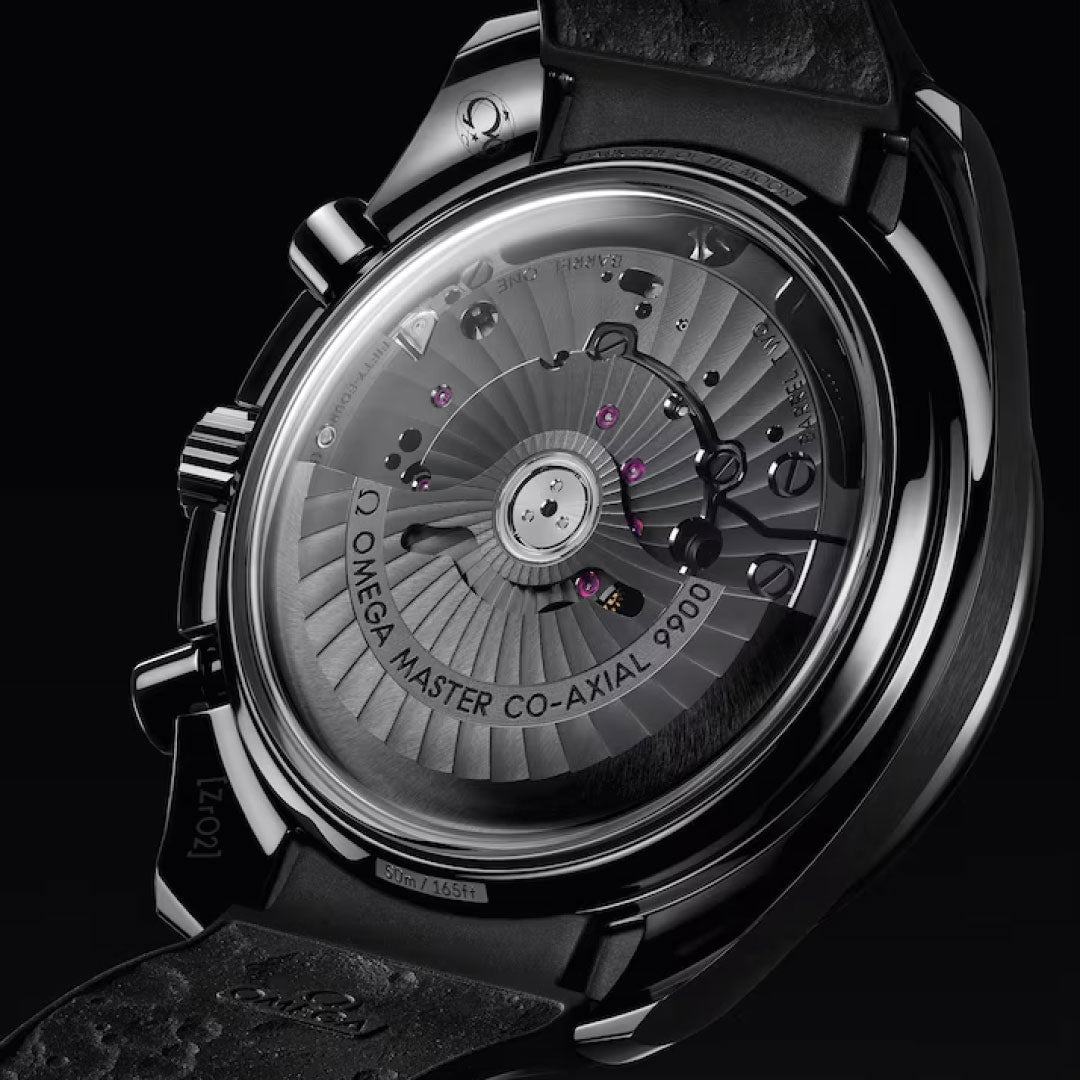 Close-up of the OMEGA Speedmaster Dark Side of the Moon Co-Axial Master Chronometer 44.25mm Watch reveals its mechanical movement and “Omega Master Co-Axial 9900” engraving through a clear case back, complemented by a black strap.