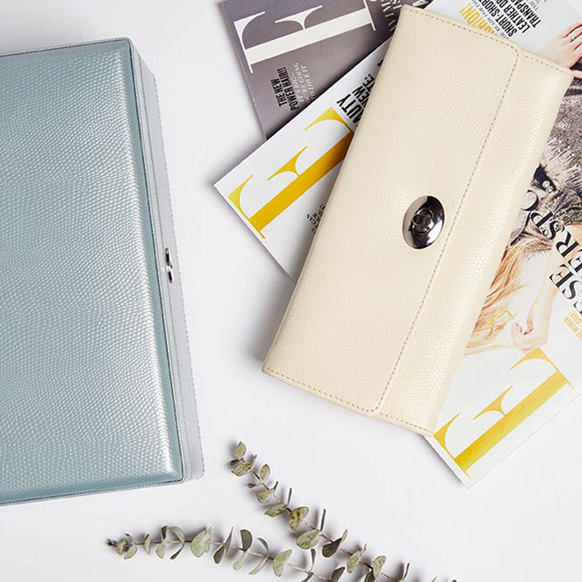A WOLF 1834 London Jewellery Roll in light blue lizard embossed leather accompanies a cream clutch with a clasp on a surface filled with fashion magazines. Small eucalyptus branches nearby add a touch of natural elegance, and the LusterLoc™ lining ensures your treasures remain pristine.