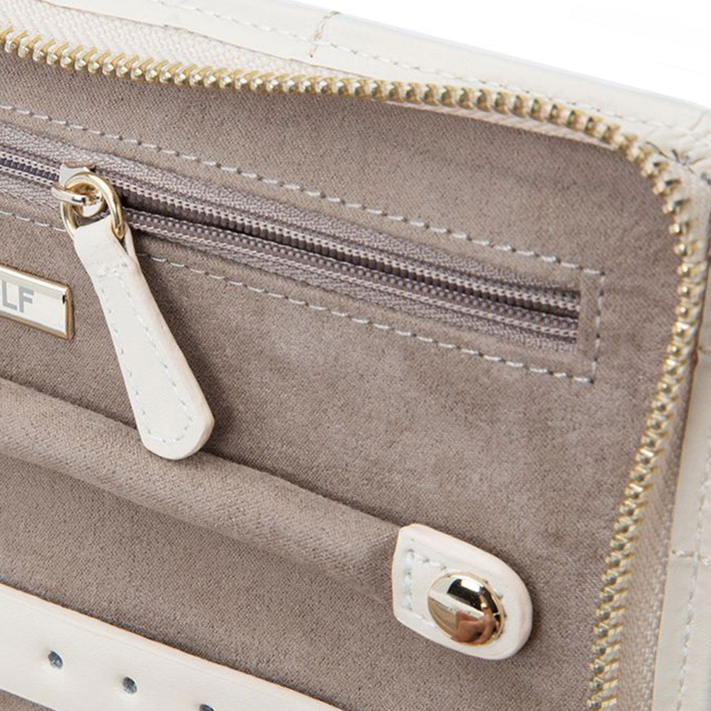 Close-up of the WOLF 1834 Caroline Jewellery Portfolio interior showcases a cream and beige color scheme with a zipper pocket, white pull tab, and gold-toned metal accents. The smooth LusterLoc™ lining complements the high-quality stitching on the diamond-quilted leather, giving it a sleek and sophisticated design.