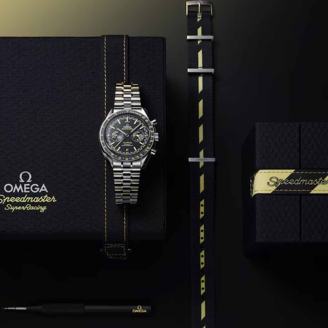 An OMEGA Speedmaster Super Racing Co-Axial Master Chronometer 44.25mm watch, featuring a metal bracelet, is elegantly displayed on its branded box. Accompanying it is an additional black strap with yellow stitching, arranged parallel to a tool and another box labeled "Speedmaster," emphasizing its Co-Axial Master Chronometer precision and innovative Spirate System technology.