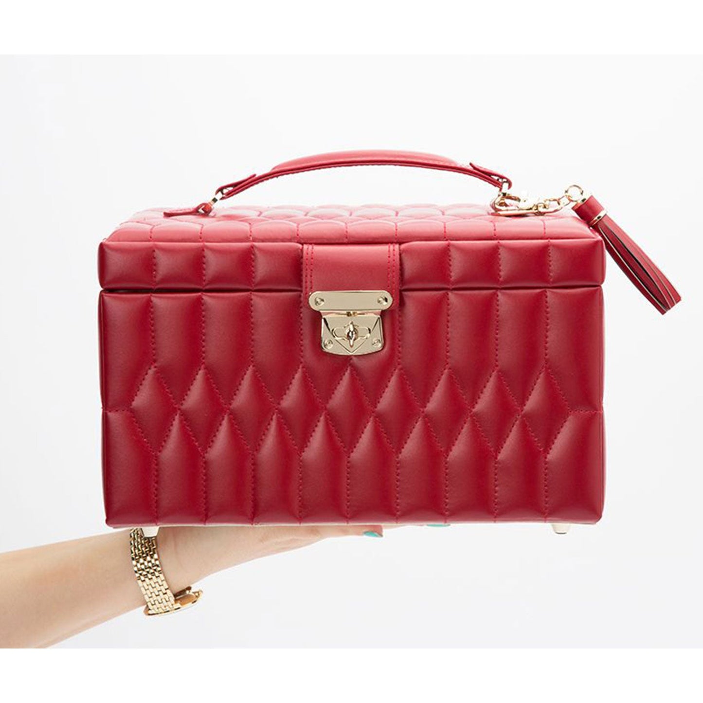 A hand holds the WOLF 1834 Caroline Red Jewellery Box, made from quilted leather with a top handle and a gold clasp in the center. With its structured rectangular design and a decorative tassel on the side, it exudes elegance akin to an impeccably styled jewellery case.