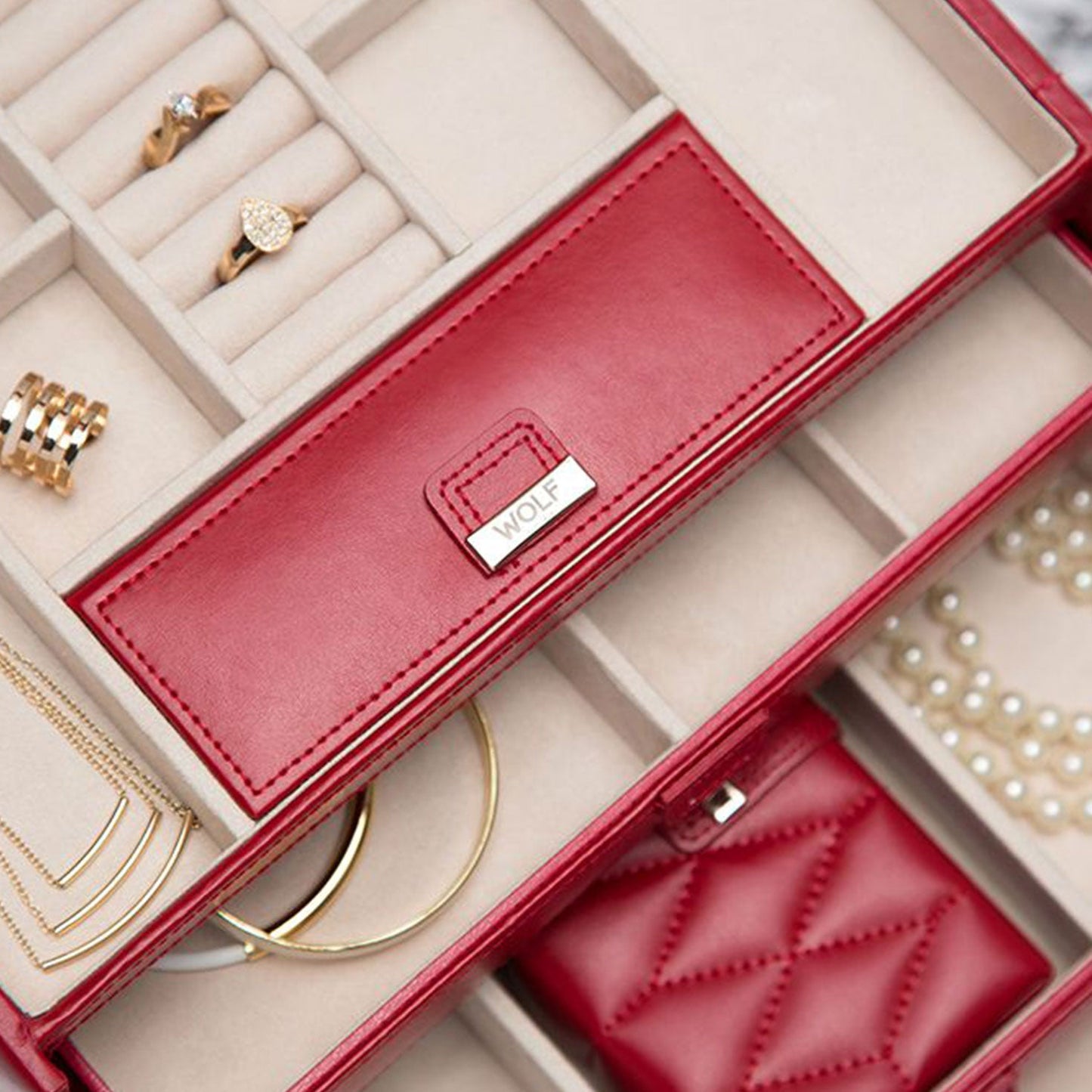 The WOLF 1834 Caroline Red Jewellery Box, crafted with diamond quilted leather, offers multiple compartments for rings, earrings, a necklace, and a bracelet. Its interior boasts cream fabric with LusterLoc™ technology, and one section features the "WOLF" label.