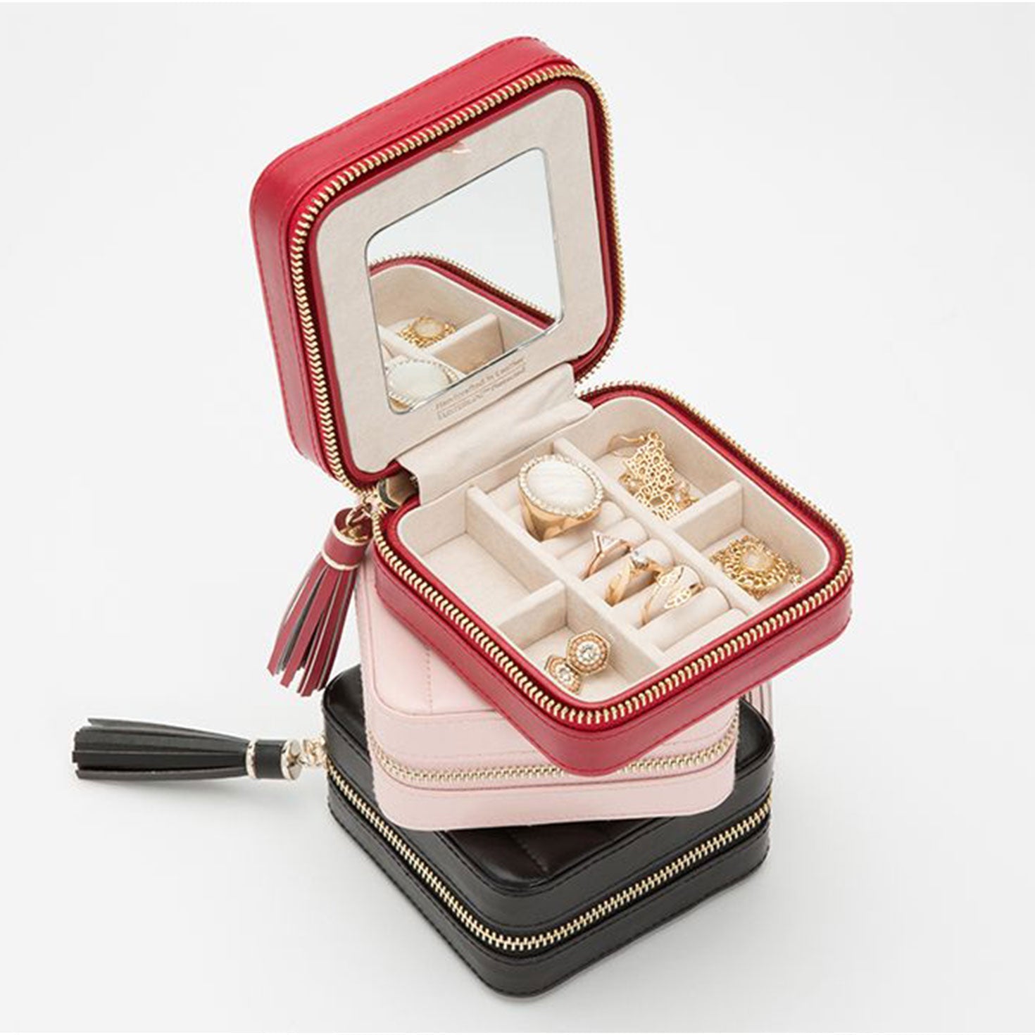 A set of three square WOLF 1834 Caroline Zip Travel Cases in red, pink, and black. The red case is shown open, displaying a mirror inside the lid and multiple compartments filled with rings and various pieces. Each case from WOLF features LusterLoc™, a zippered closure, tassel accents, and diamond-quilted leather design.