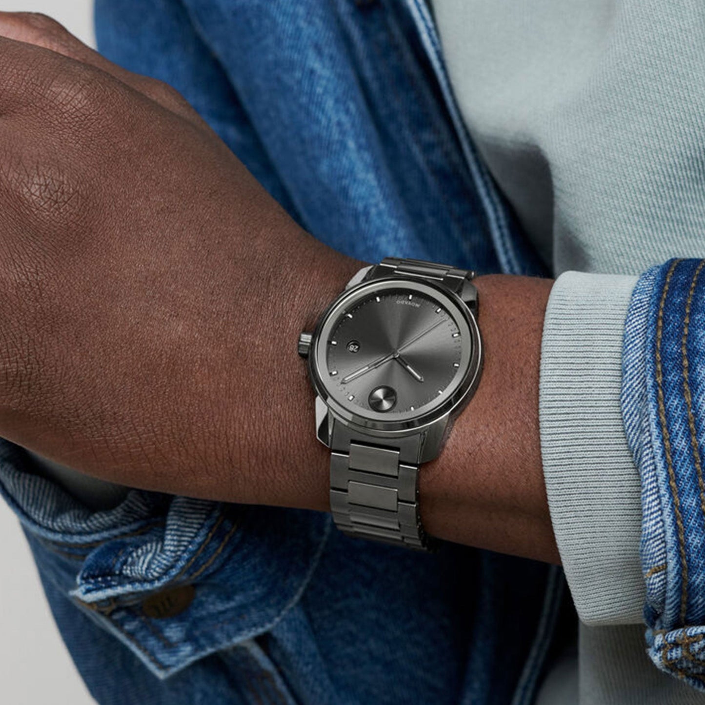 A person is casually crossing their arm, showcasing a Movado BOLD Verso Quartz 42mm Watch, designed with a sleek silver metallic band. Their light gray sweater is visible beneath a denim jacket. The watch face maintains a modern and minimalist design, enhanced by Swiss Super-LumiNova® technology for exceptional clarity.