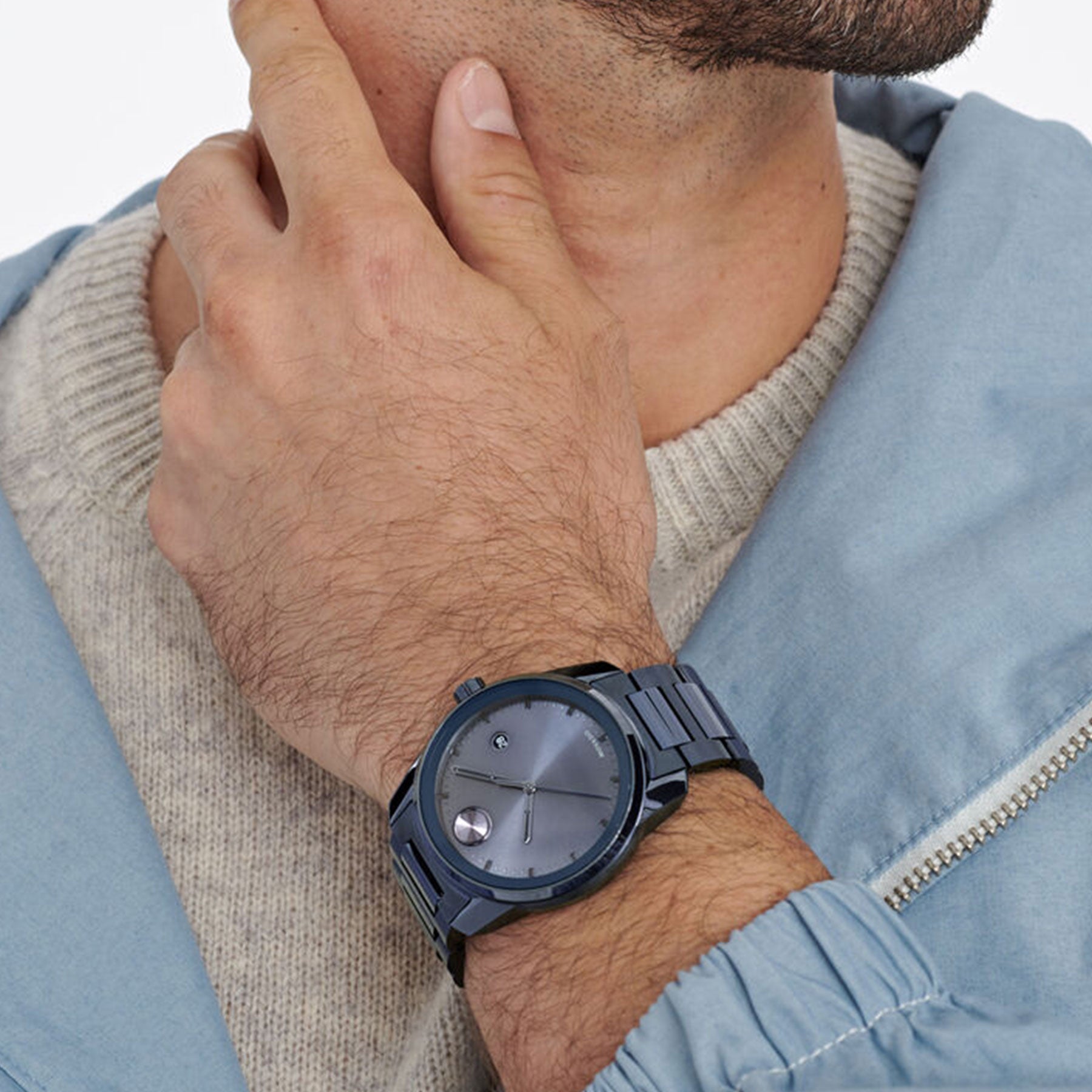 A person wearing a light blue jacket and beige sweater poses thoughtfully with their hand on their chin. On their left wrist, they sport the Movado BOLD Verso Quartz 42mm Watch by Movado. This sleek, dark blue timepiece features a minimalist design and Swiss Super-LumiNova® glow, perfectly complementing their stylish ensemble.