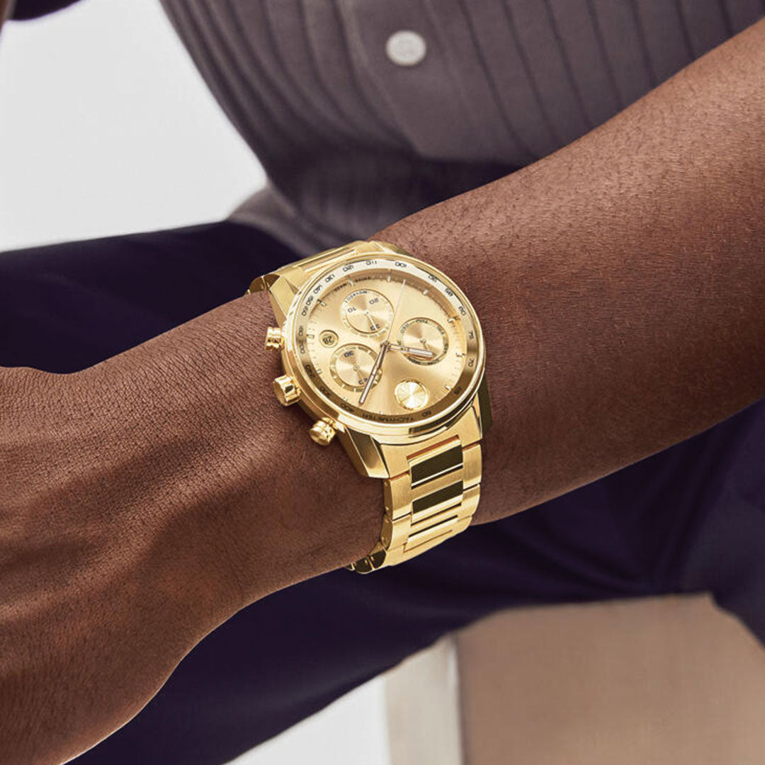 A close-up shows a person’s wrist wearing the Movado BOLD Verso Chronograph Quartz 44mm Watch with a gold band, set against dark pants and a pinstriped shirt.