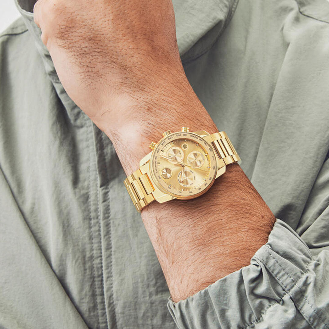 A person in a light gray long-sleeve jacket showcases the Movado BOLD Verso Chronograph Quartz 44mm watch with a yellow gold stainless steel case, multiple subdials, and a tachymeter bezel on their left wrist.