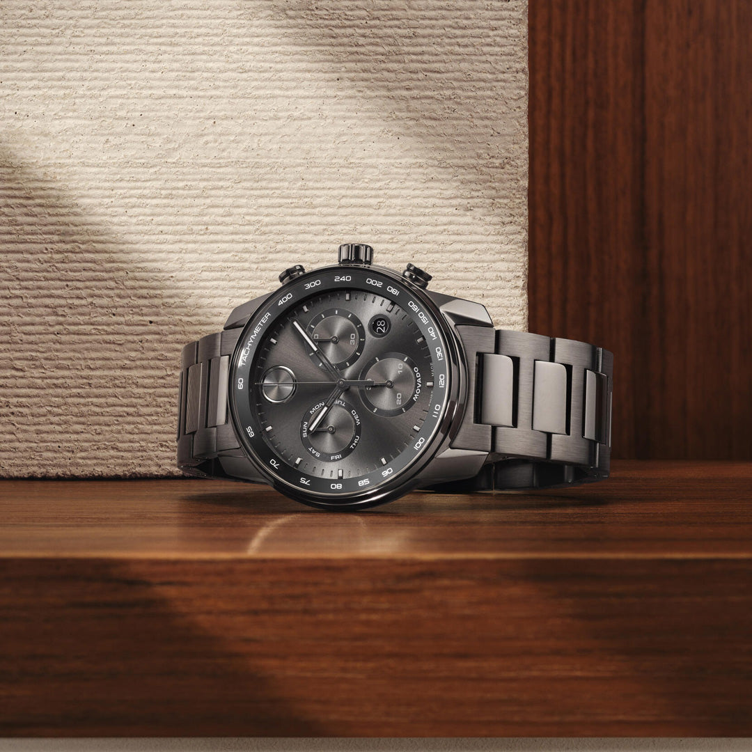 The Movado BOLD Verso Chronograph Quartz 44mm Watch by Movado, featuring a silver-tone metal design, a dark dial with three subdials, and a tachymeter bezel, rests on a wooden surface beside a textured beige wall.
