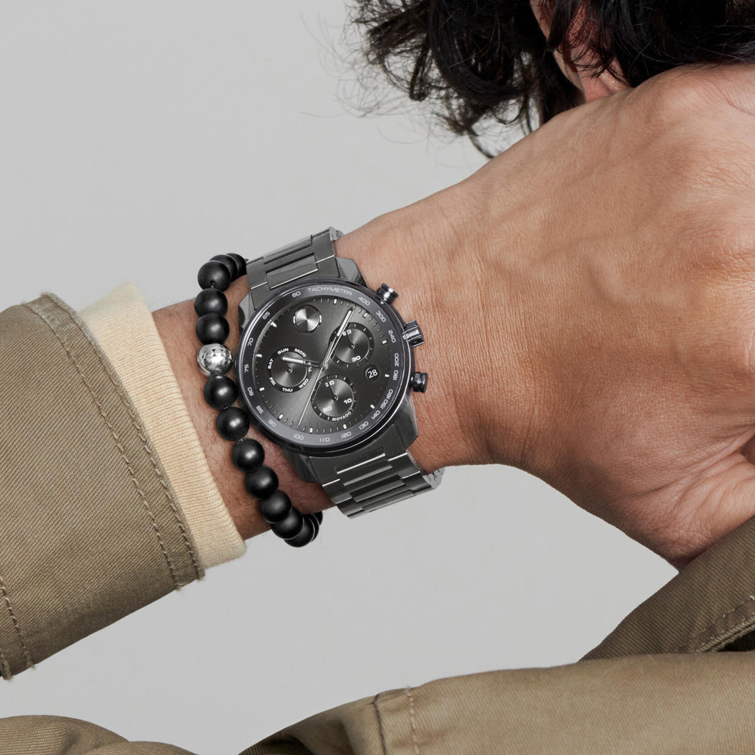 A person wearing the Movado BOLD Verso Chronograph Quartz 44mm Watch by Movado, featuring a silver case and tachymeter bezel, paired with a black beaded bracelet accented by a skull bead on their left wrist, dressed in a beige jacket.