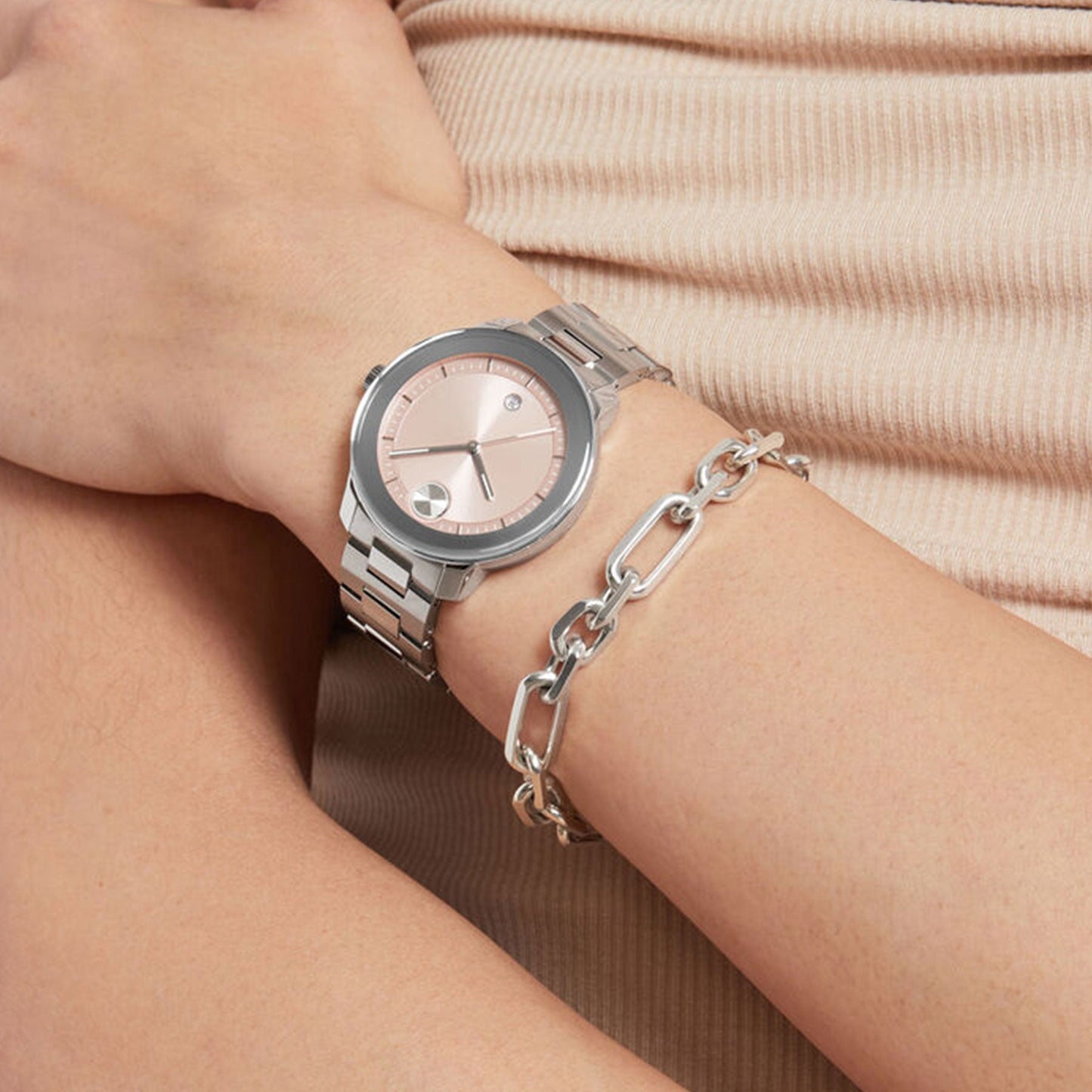 A close-up of a person sporting the Movado BOLD Verso Quartz 38mm, a contemporary-chic watch in stainless steel with a blush dial on their left wrist. Next to the Movado watch is a silver link bracelet. The person's arm rests on their lap, clad in beige ribbed fabric.