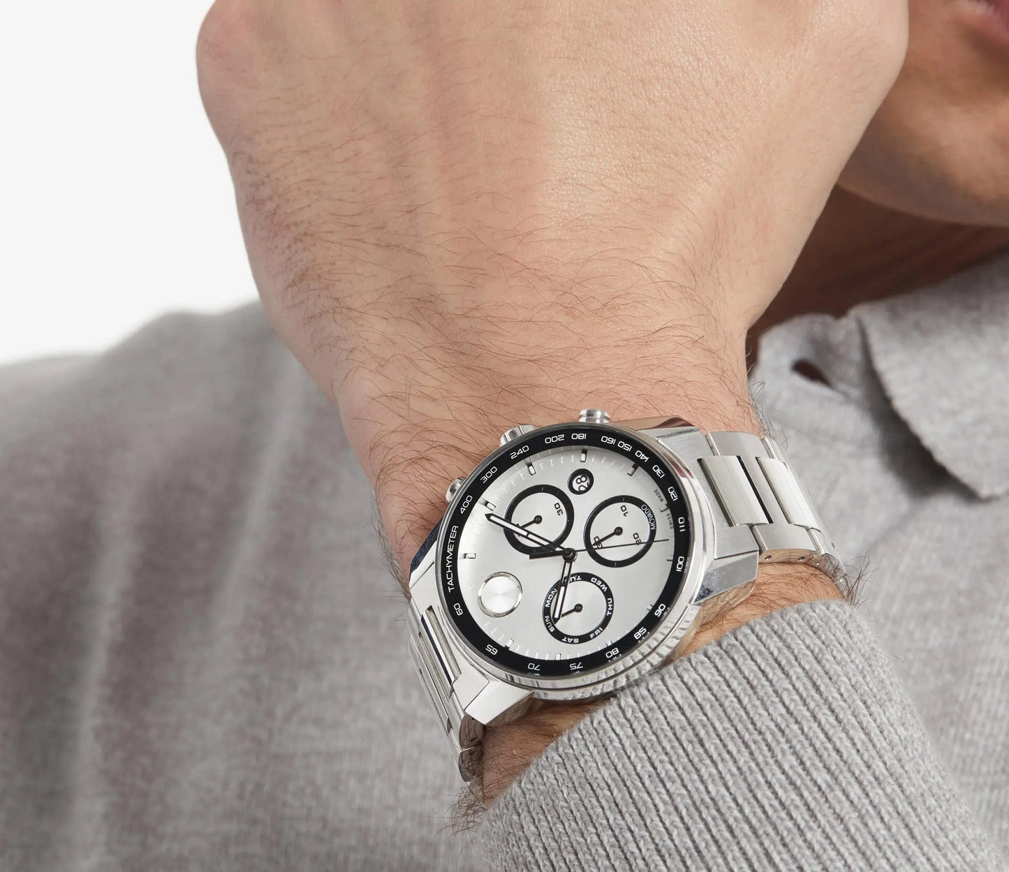 A person wears a Movado BOLD Verso Quartz 44mm Watch, showcasing a stainless steel design with a white and black chronograph dial surrounded by a tachymeter bezel. The look is completed with a light gray, long-sleeved shirt.