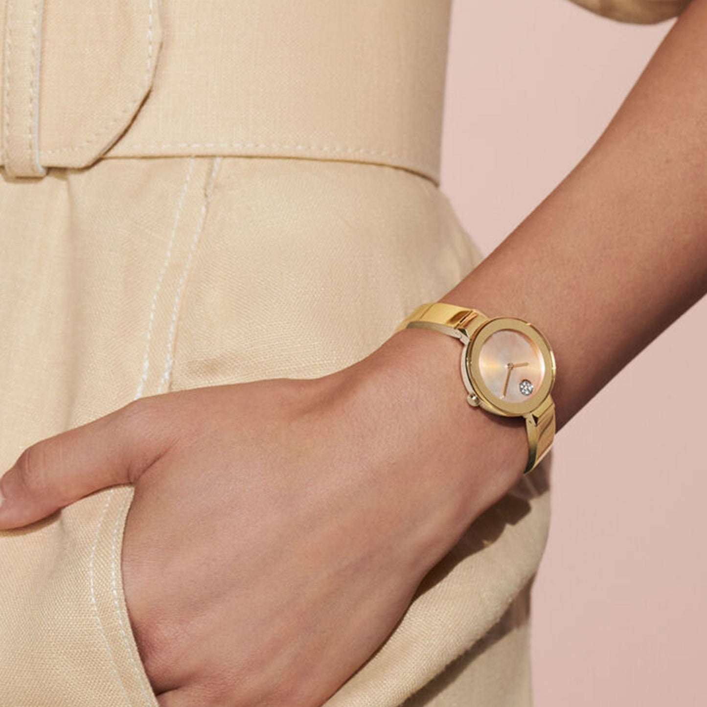 A person dressed in a beige ensemble with one hand in their pocket is displaying a Movado BOLD Quartz 28mm Watch on their wrist. The watch, by Movado, boasts a minimalistic face and features a mother-of-pearl dial adorned with a small diamond accent. The backdrop consists of soft, neutral tones.