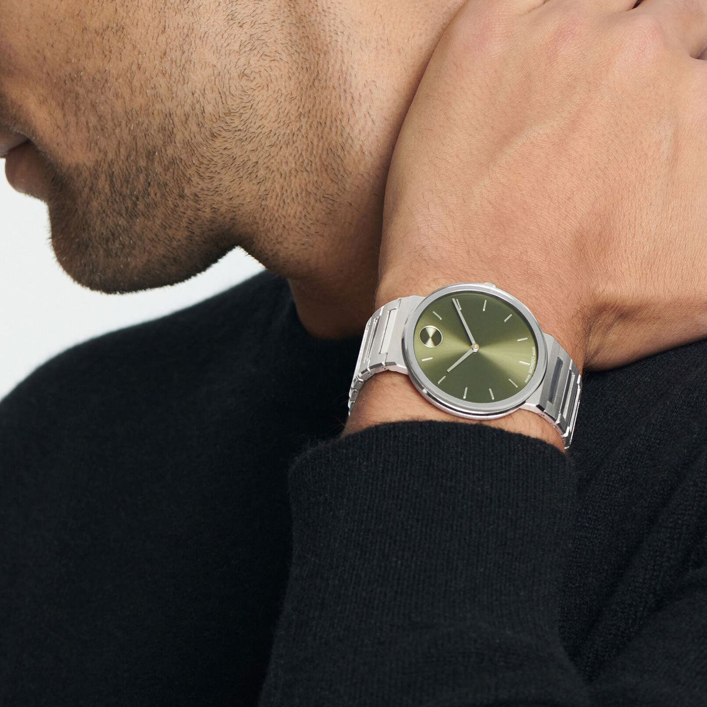 A person sports a black sweater, showcasing the Movado BOLD Horizon Quartz 40mm Watch with a minimalist silver design and green face on their left wrist. Their right hand is casually resting on their neck, creating an effortlessly stylish look.