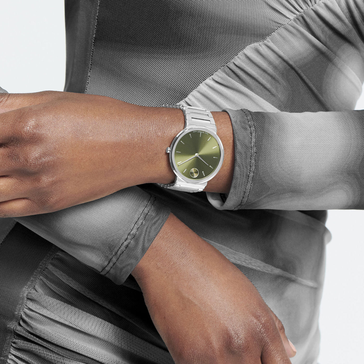 A person wearing a sleek gray dress showcases the Movado BOLD Horizon Quartz 40mm Watch, featuring a shimmering green face and silver strap. The dress is designed with a textured, ruched pattern, enhancing their sophisticated allure.