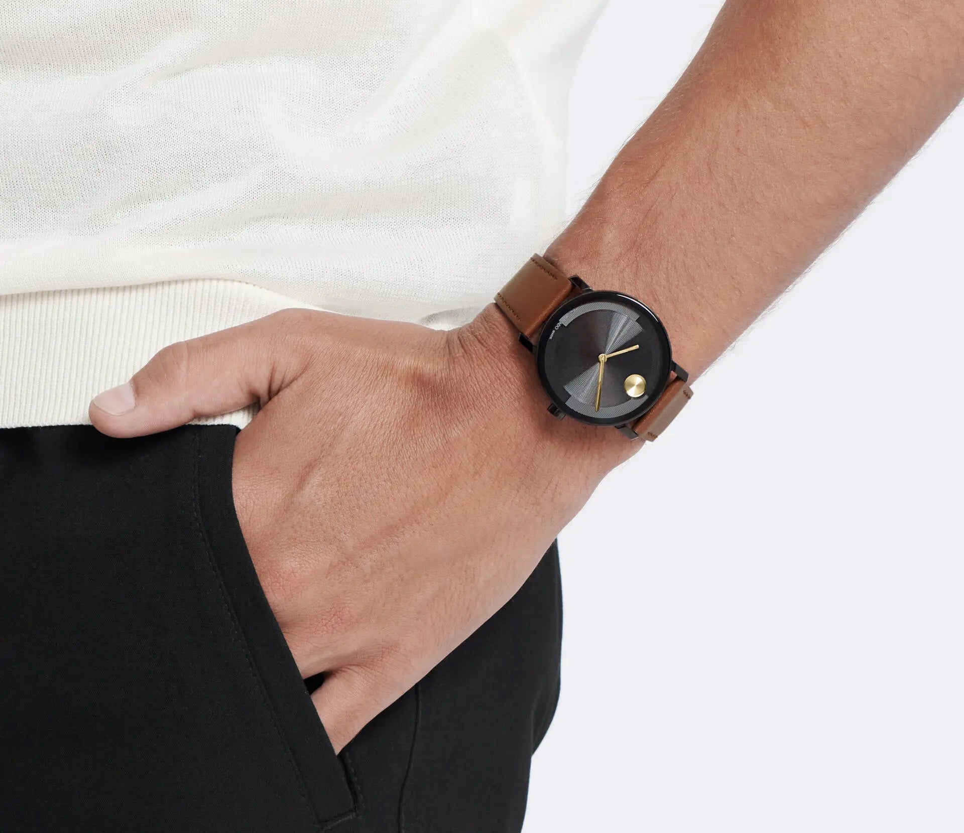 A person wearing a white shirt and black pants has their hand in their pocket, showcasing the Movado BOLD Evolution 2.0 Quartz 40mm Watch. This stylish timepiece features a black face, golden hands, and a luxurious cognac leather strap. The background is plain white.