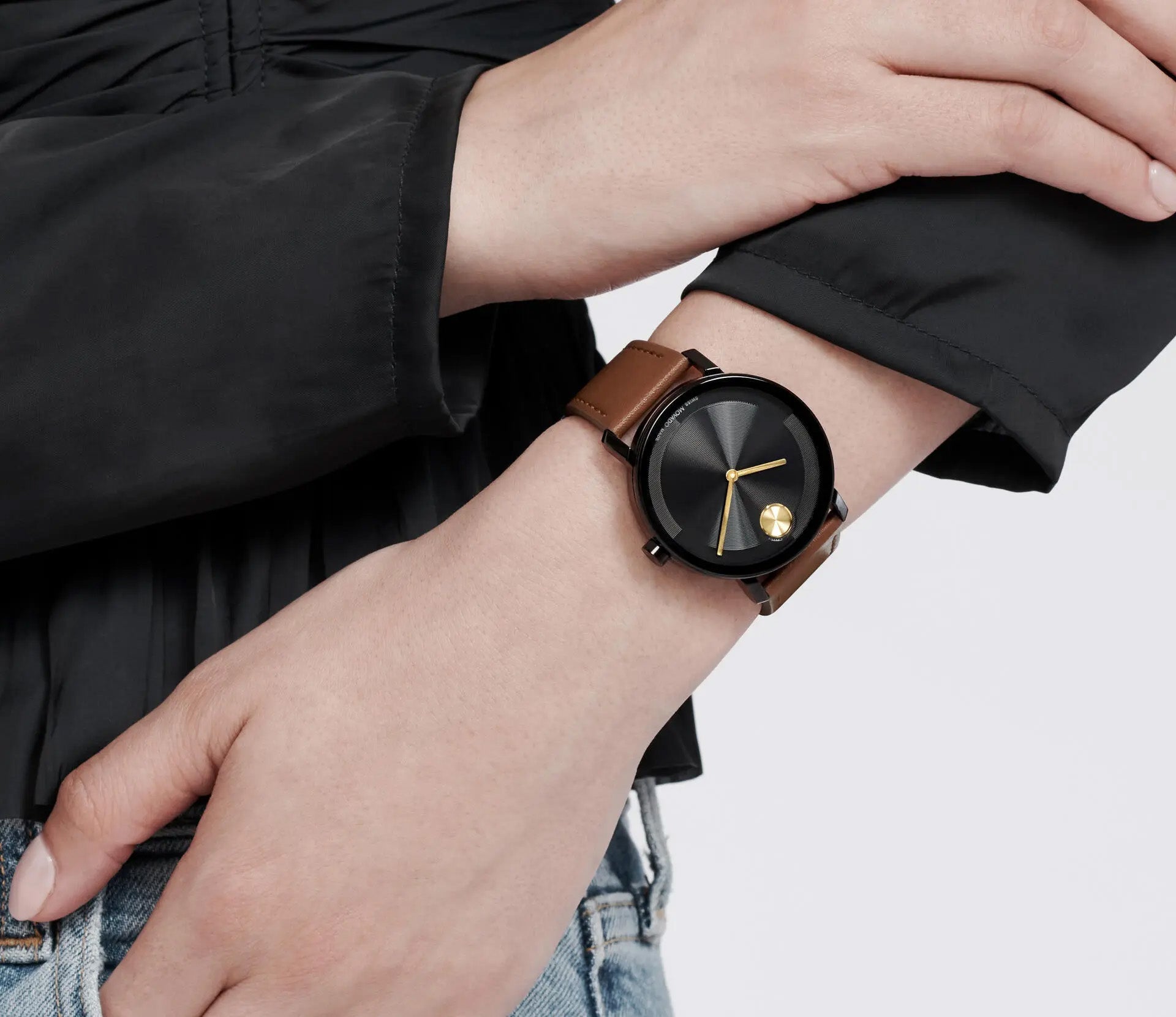 A person wears the Movado BOLD Evolution 2.0 Quartz 40mm Watch, featuring a rich brown cognac leather strap, a black face with gold hands, and a micro-textured tonal dial. The watch graces their wrist as they sport a black shirt and blue jeans against a plain white background.