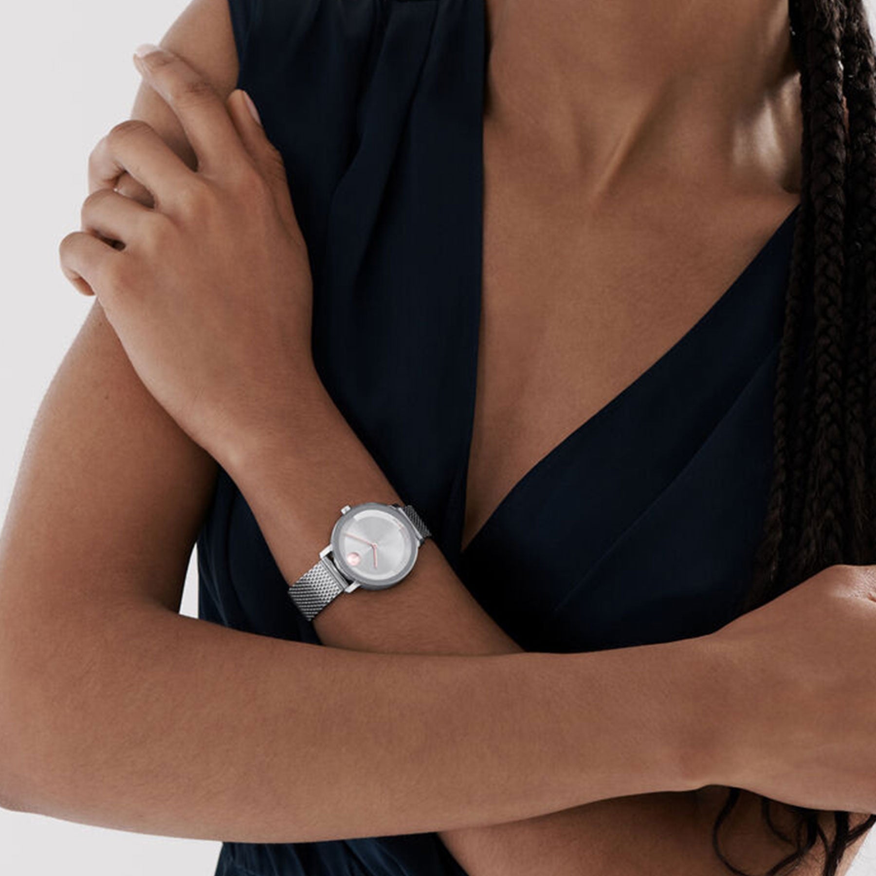 Dressed in a dark, sleeveless top, a person crosses their arms to highlight the minimalist and sophisticated Movado Amorosa Quartz 24mm Watch on their wrist. The watch's sleek design and simple face add elegance to the ensemble. A plain, light-colored background further enhances this stylish look.