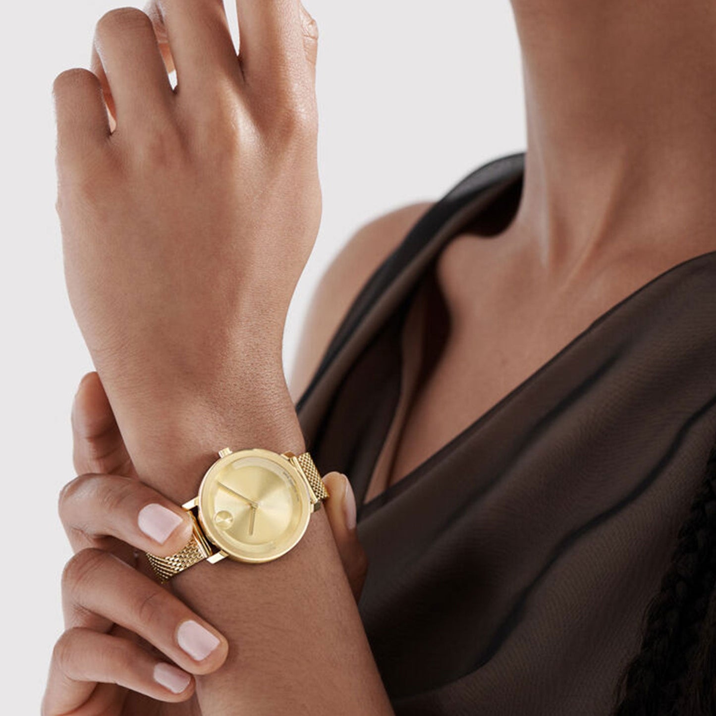 A person wearing a sleeveless black top showcases the elegant Movado BOLD Evolution Quartz 34mm Watch, complete with a stylish goldtone mesh band that captures the essence of minimalist design. Featuring light pink nail polish, they hold their right hand near their face against a simple, light gray background.