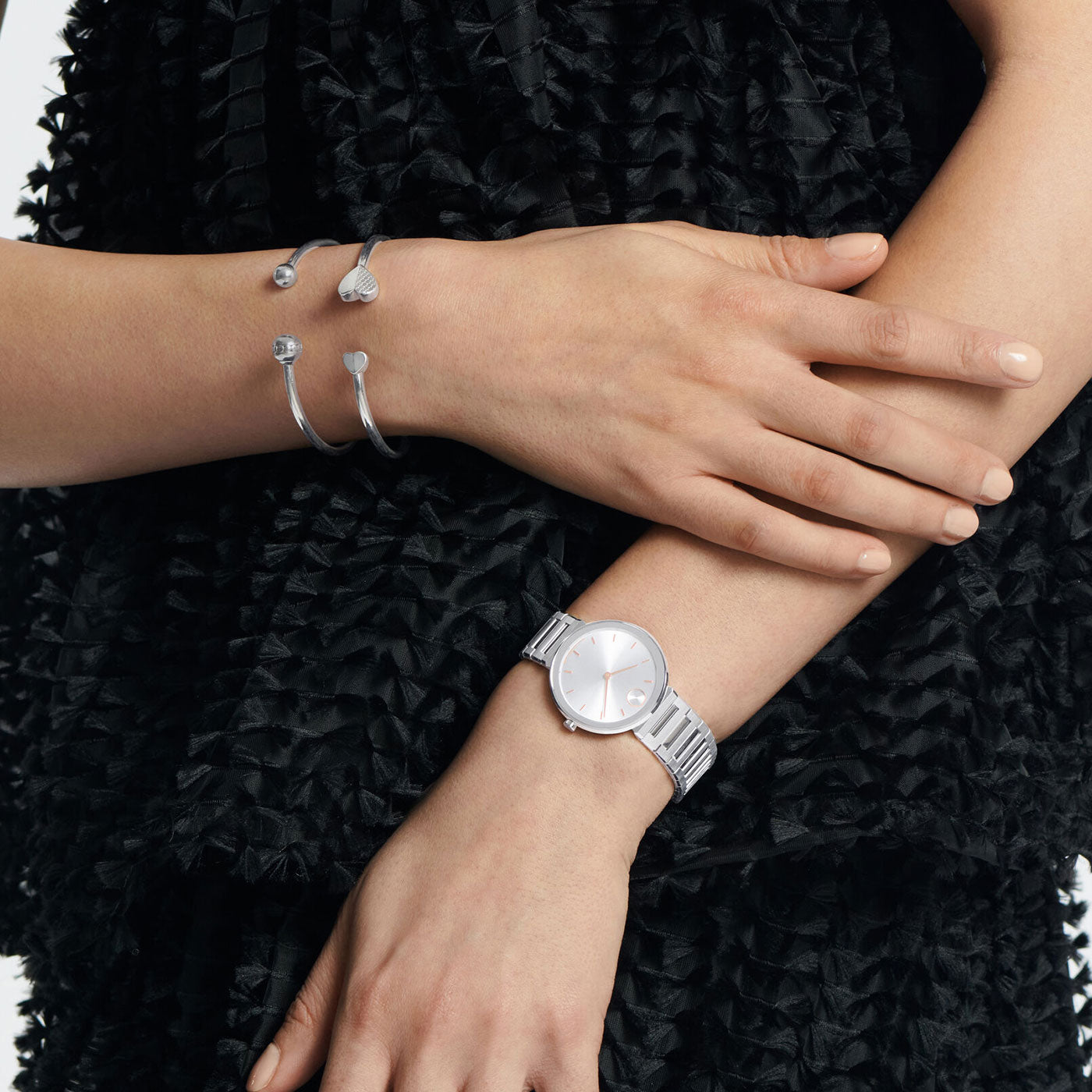 A person in a black textured dress crosses their arms, showcasing the elegant Movado BOLD Horizon Quartz 34mm Watch and two stylish silver bangles on one wrist. The watch features a simple round face and a metal link band.