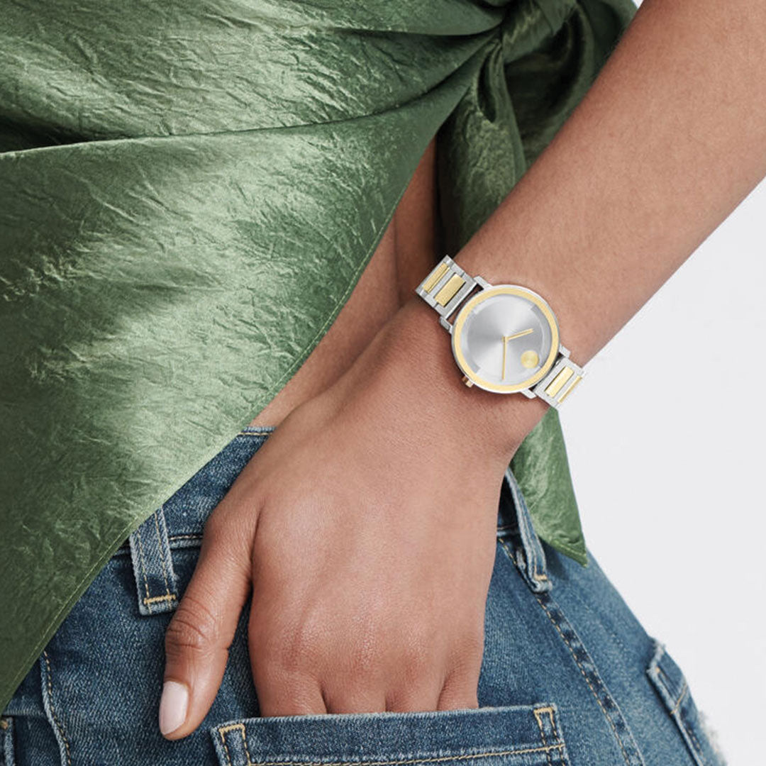 A person in blue jeans and a green textured top rests their hand on their hip, highlighting the Movado BOLD Evolution 2.0 Quartz 34mm Watch with a silver and goldtone bracelet.