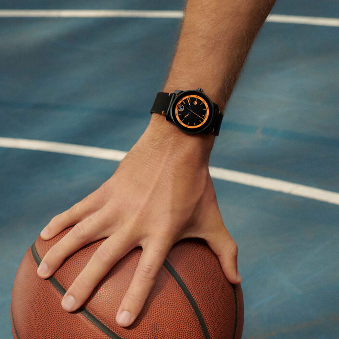 A hand wearing a Movado BOLD TR90 Quartz 40mm Watch, featuring an orange-accented face and black leather strap, rests on a basketball. In the background, the blue surface of a basketball court is illuminated by the Swiss Super-LumiNova glow against the stark white line.