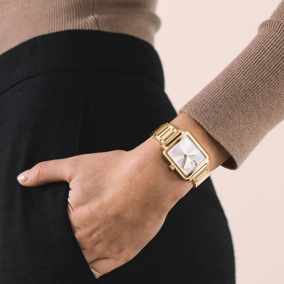 A person wearing the Movado BOLD Evolution 2.0 Quartz 28mm x 26mm Watch with a light blue lacquer dial and a beige long sleeve top has their hand in the pocket of black pants against a neutral background.