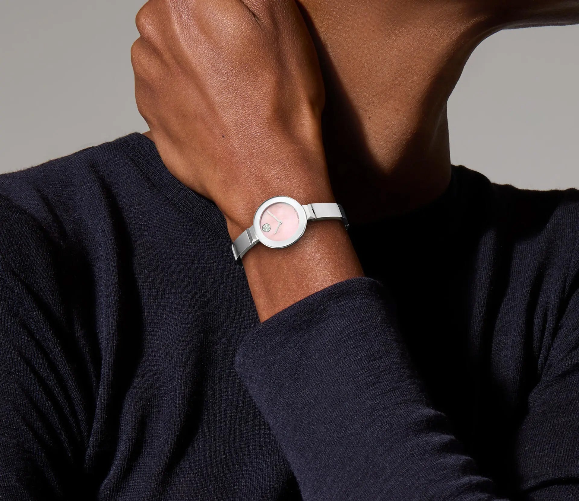 A person with their hand on their chin is accessorized with the Movado BOLD Bangle Quartz 28mm Watch, featuring a minimalist design with a light pink face and silver band that subtly resembles a Mother-of-Pearl finish. They are wearing a dark, long-sleeve top against a neutral background.