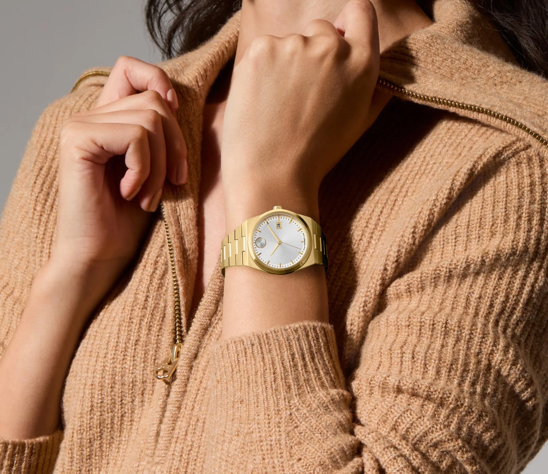 A person wearing a beige knit sweater is seen adjusting the zipper, and on their wrist is prominently displayed the Movado BOLD Quest Quartz 35mm Watch. The background is a soft, neutral color.