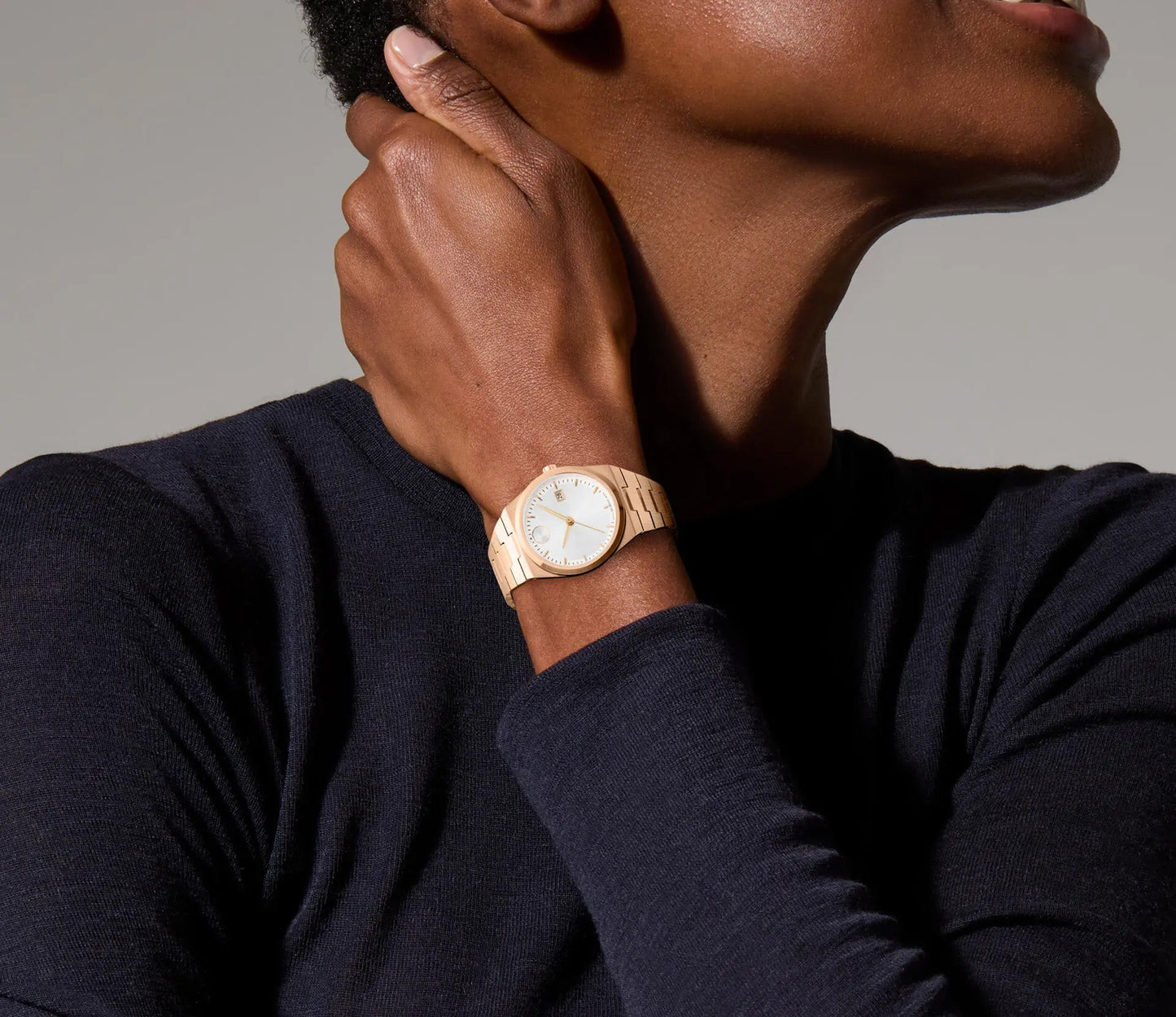 A person wearing a dark shirt holds their hand near their face, highlighting the Movado BOLD Quest Quartz 35mm Watch. The watch's gold wristband and round face are elegantly complemented by rose gold tone hands, emphasizing its minimalist design.