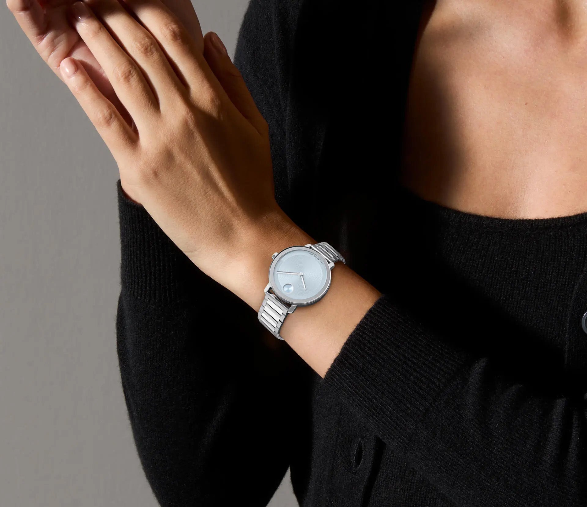 A person wearing the Movado BOLD Evolution 2.0 Quartz 34mm Watch with an ice blue lacquer dial adjusts their sleeve. They are dressed in a black sweater, and the background is a neutral color. The watch features a simple, minimalist design.