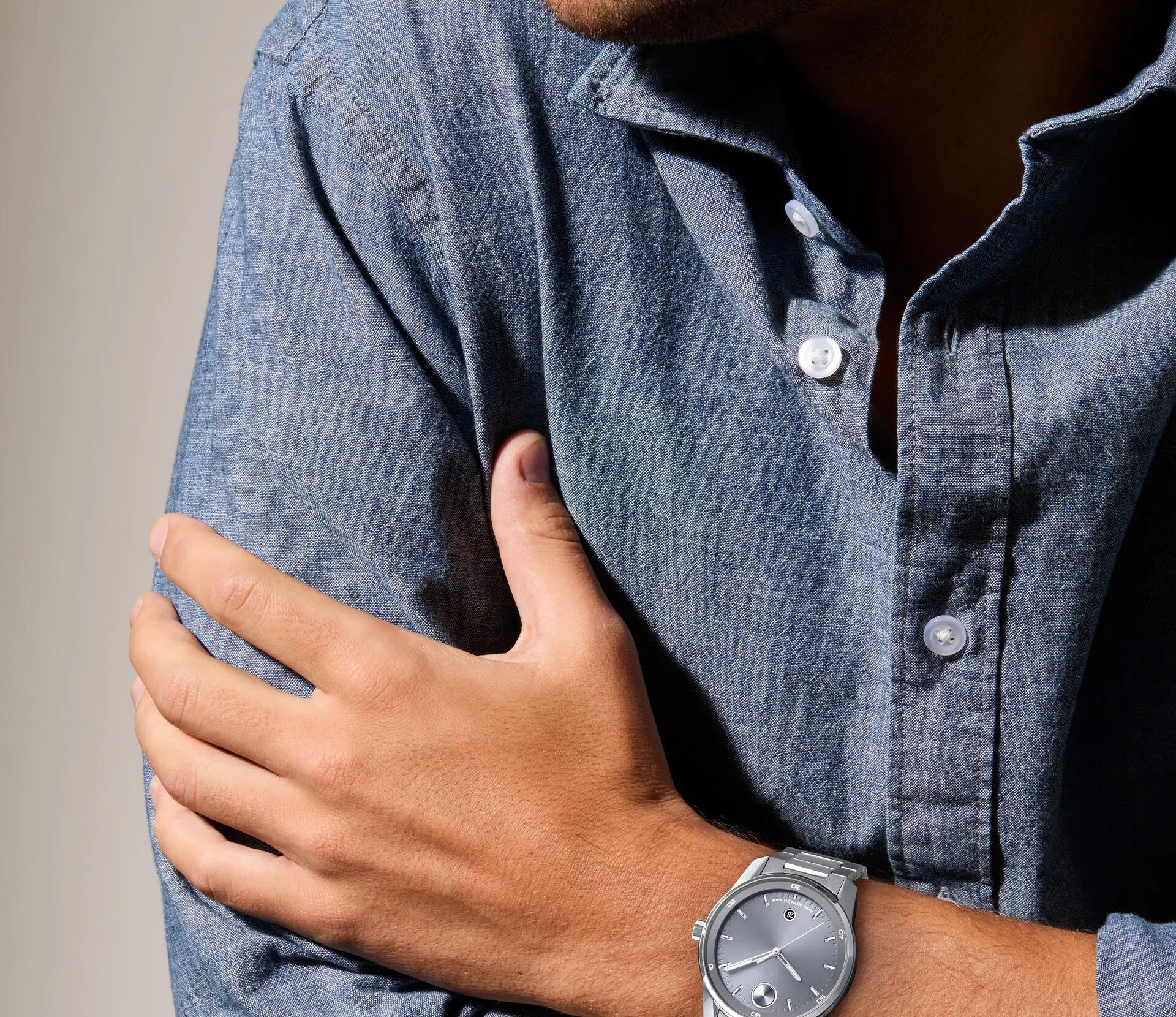 A person wearing a bold blue denim long-sleeve shirt and a silver Movado BOLD Verso Quartz 42mm watch, featuring Swiss Super-LumiNova, is folding their arms. The sport-inspired watch from Movado showcases a grey face that stands out prominently in the image.