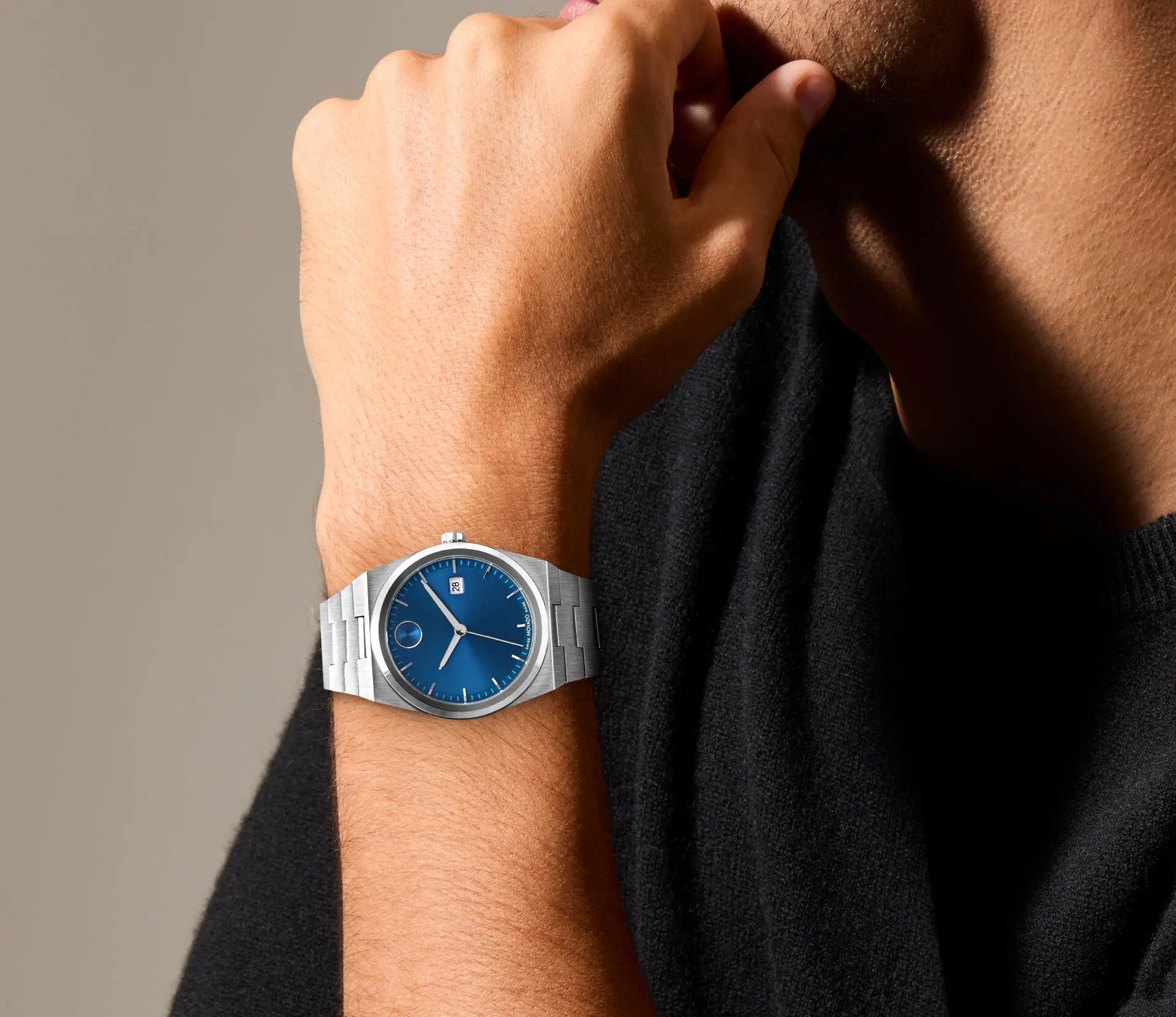 A person wearing the Movado BOLD Quest Quartz 40mm Watch with its striking blue sunray dial and date display casually rests their chin on their hand. This silver wristwatch, featuring an integrated bracelet, effortlessly complements a black shirt, creating a sophisticated yet bold look.