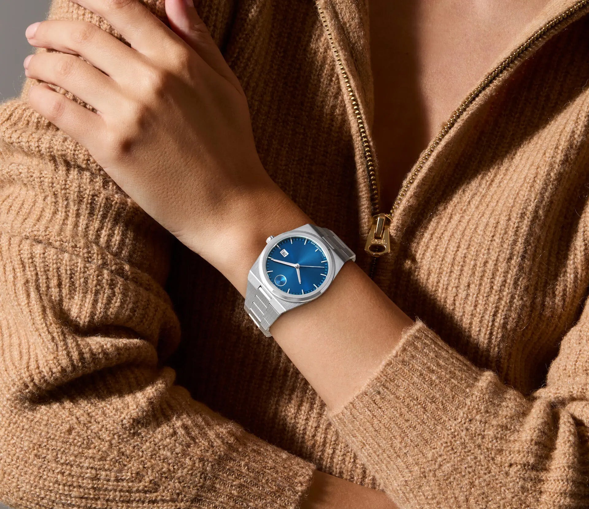 A person wearing a tan sweater with a zipper partially open, revealing the striking blue sunray dial of the Movado BOLD Quest Quartz 40mm Watch. With their left hand resting on their right arm, they proudly showcase this elegant timepiece from Movado.