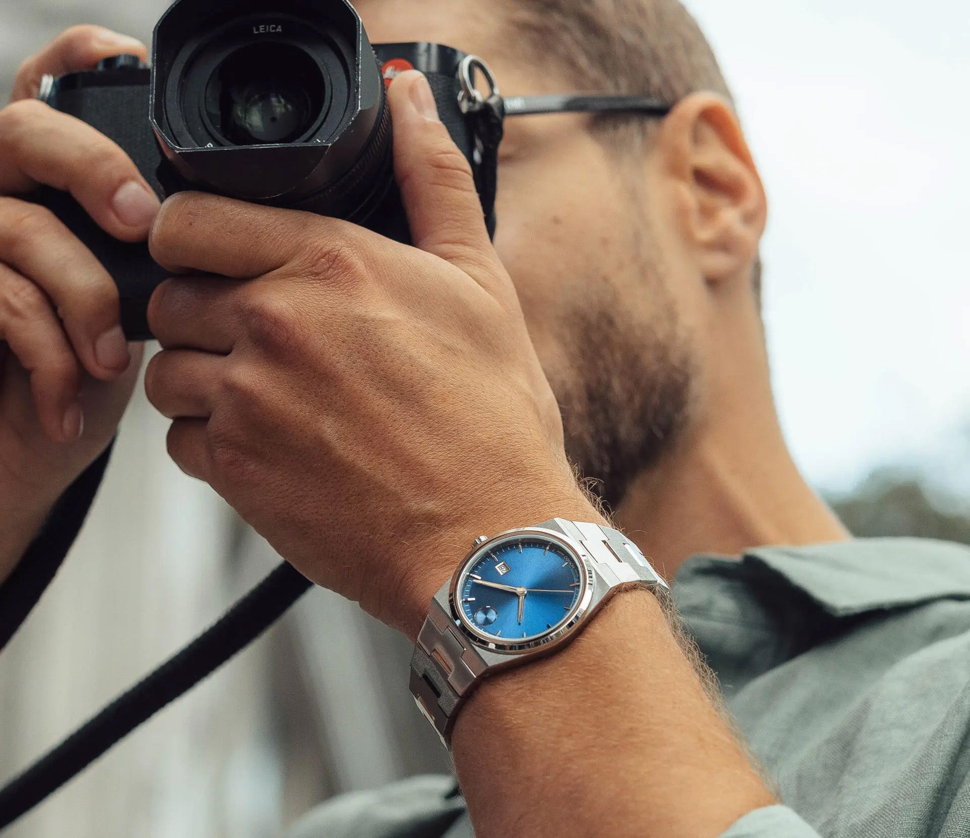 A person wearing a green shirt is holding a camera up to their face, capturing a photo. A close-up of their wrist reveals the Movado BOLD Quest Quartz 40mm Watch, featuring an integrated silver bracelet and a striking blue sunray dial.