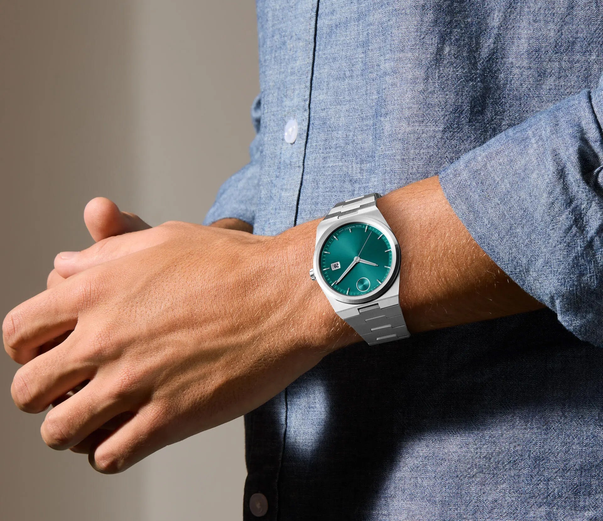 A person wearing a light blue shirt showcases the Movado BOLD Quest Quartz 40mm Watch with an integrated silver bracelet. The watch, featuring a green sunray dial and date display, beautifully complements the metal band. Their hands are positioned together in front of a neutral background, capturing the essence of this exquisite timepiece by Movado.