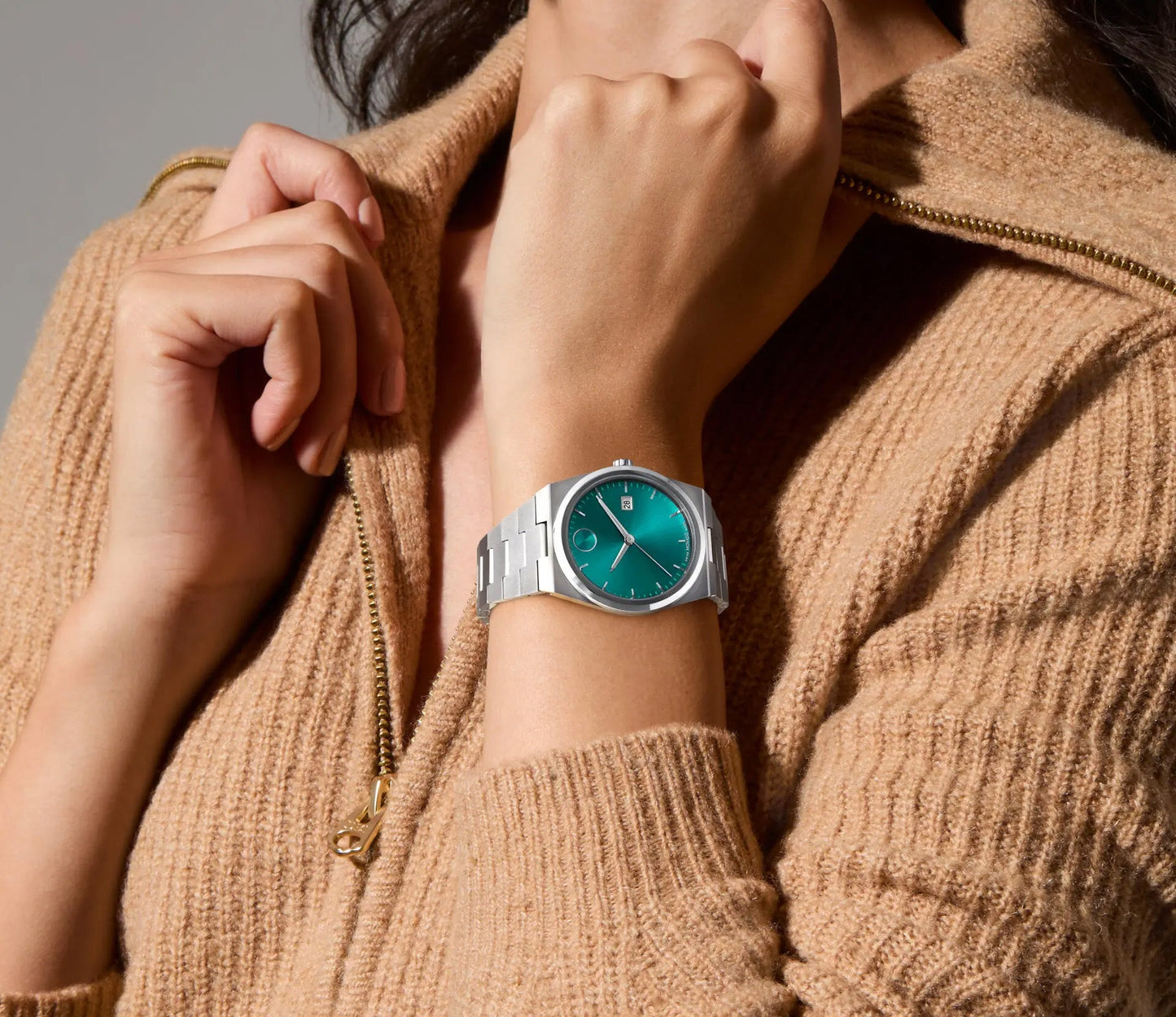 Wearing a tan zip-up sweater, an individual displays a Movado BOLD Quest Quartz 40mm Watch, featuring a striking silver wristwatch with a green sunray dial and silver hour markers that emphasize its design. The watch's integrated bracelet adds to its elegance as the person's hand rests near their chin.