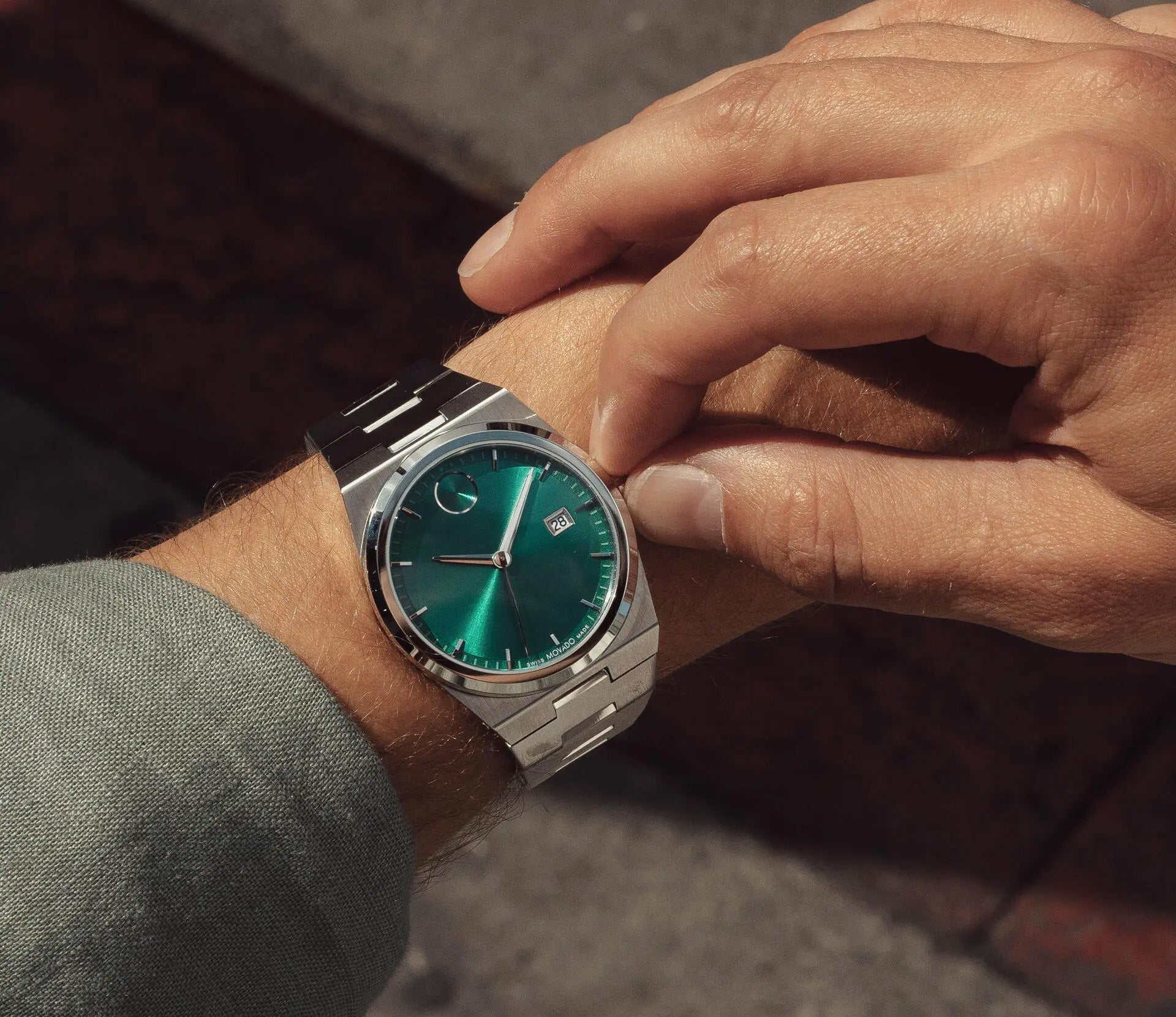 A person adjusts the Movado BOLD Quest Quartz 40mm watch, a sleek silver timepiece with an integrated bracelet and a vibrant green sunray dial featuring a date display. The watch is worn over a light green sleeve, set against the backdrop of a blurred outdoor setting.