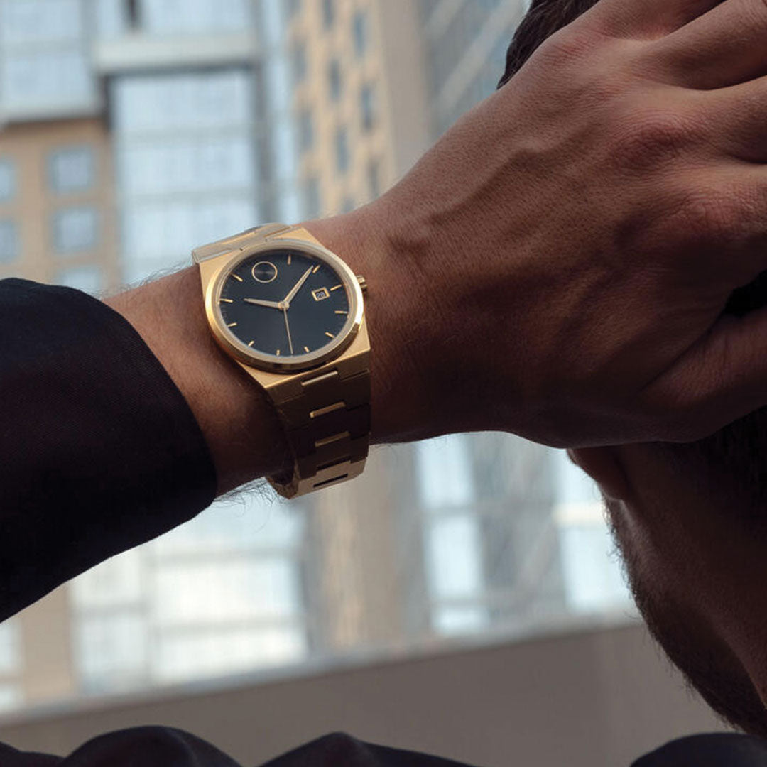Wearing a Movado BOLD Quest Quartz 40mm Watch with an integrated bracelet and black sunray dial, a person raises their hand to their head as tall, windowed buildings blur in the background.